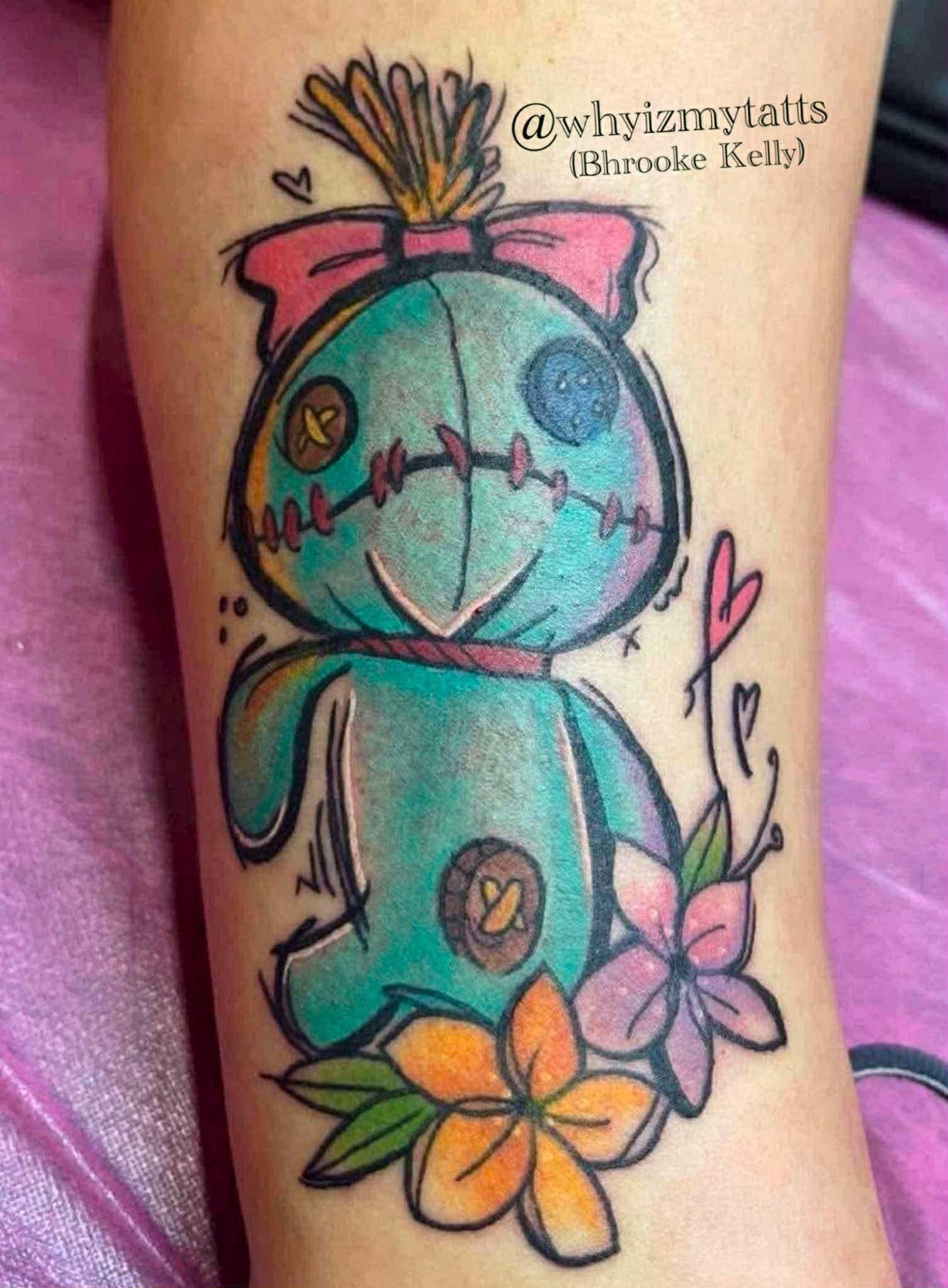 Colorful tattoo of a cartoon-style alarm clock with a pink bow on top, surrounded by pink and orange flower blossoms with green leaves, and a small heart and doodle details.