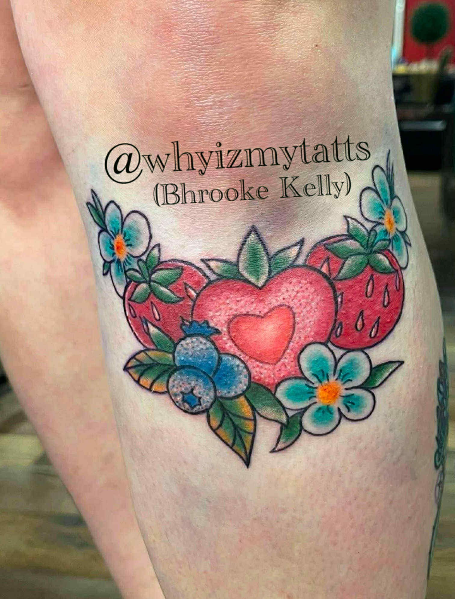 Tattoo of strawberries, blueberries, and flowers with a heart on a person's skin, with the text '@whyizmytatts (Bhrooke Kelly)'