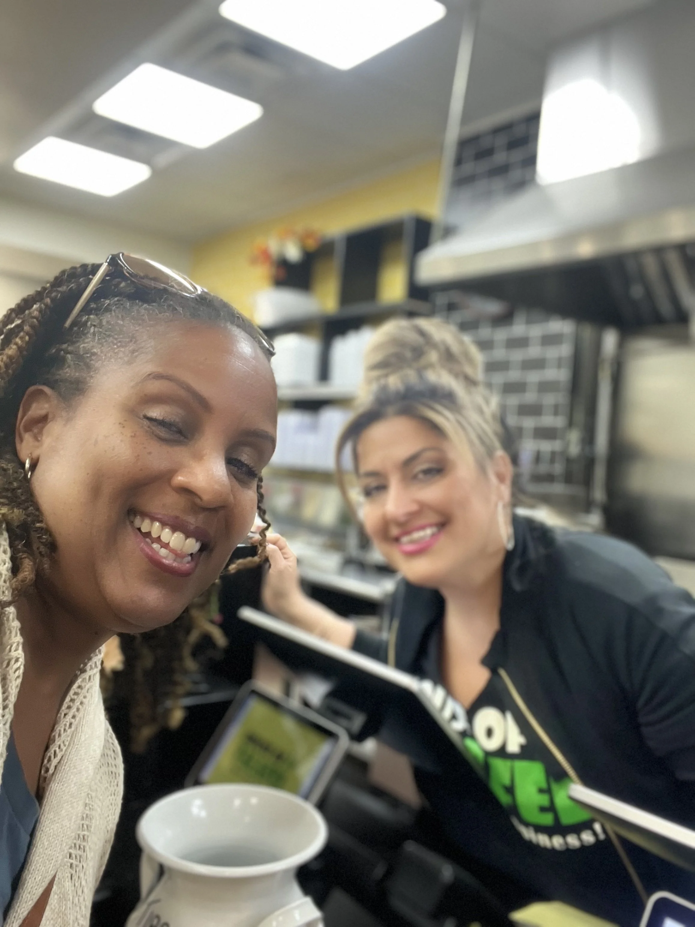 Two smiling women taking a selfie in a store or cafe, with shelves and a menu visible in the background, one holding a tablet, and a coffee mug in the foreground.