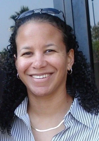 Smiling woman with curly hair, wearing glasses on top of her head, hoop earrings, a striped shirt, and a necklace, standing outside near a building with greenery in the background.