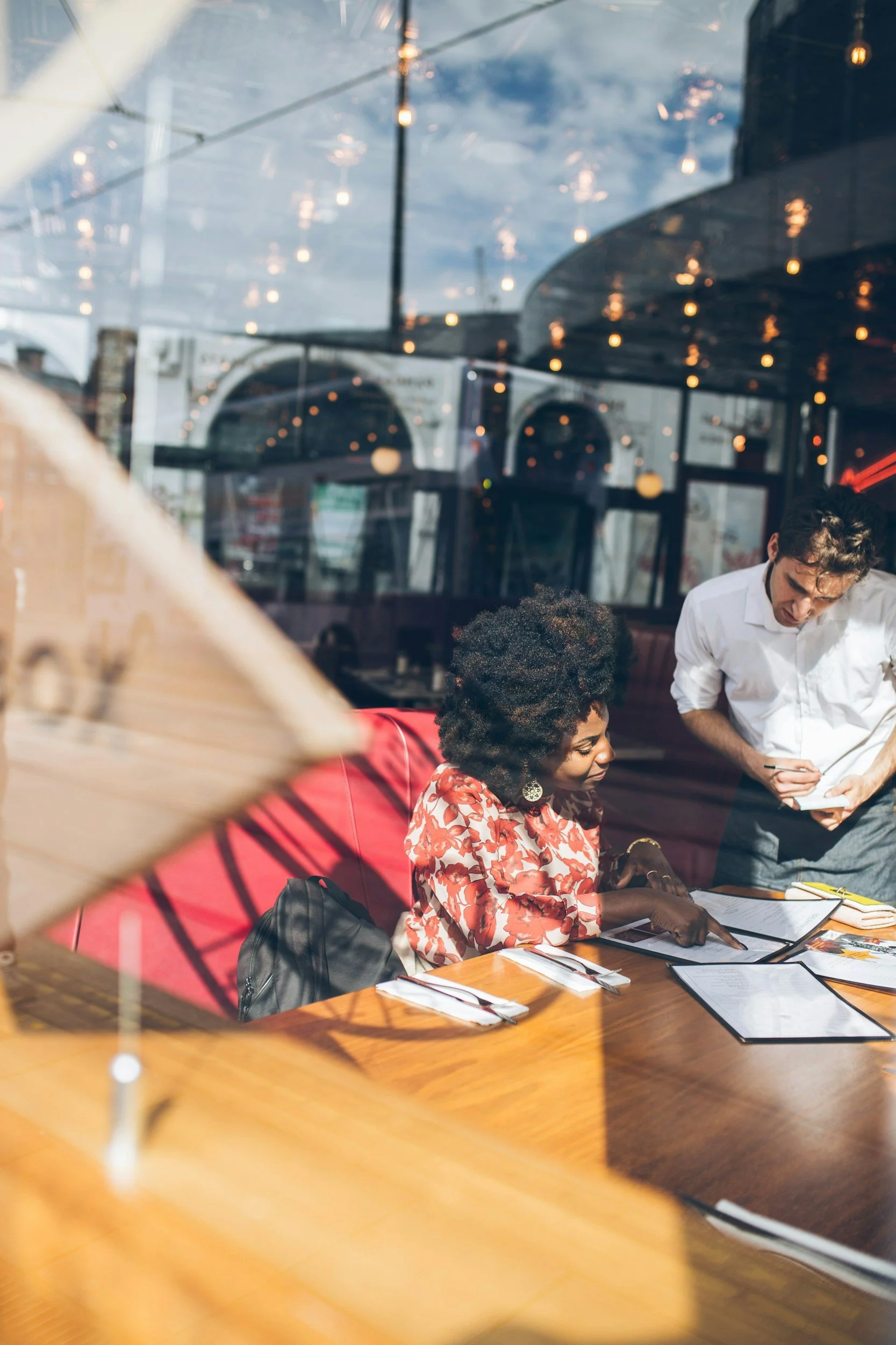 A woman with curly hair and earrings seated at a restaurant table, pointing at a menu, with a man standing next to her looking at the menu, viewed through a window with reflections of lights and sky.