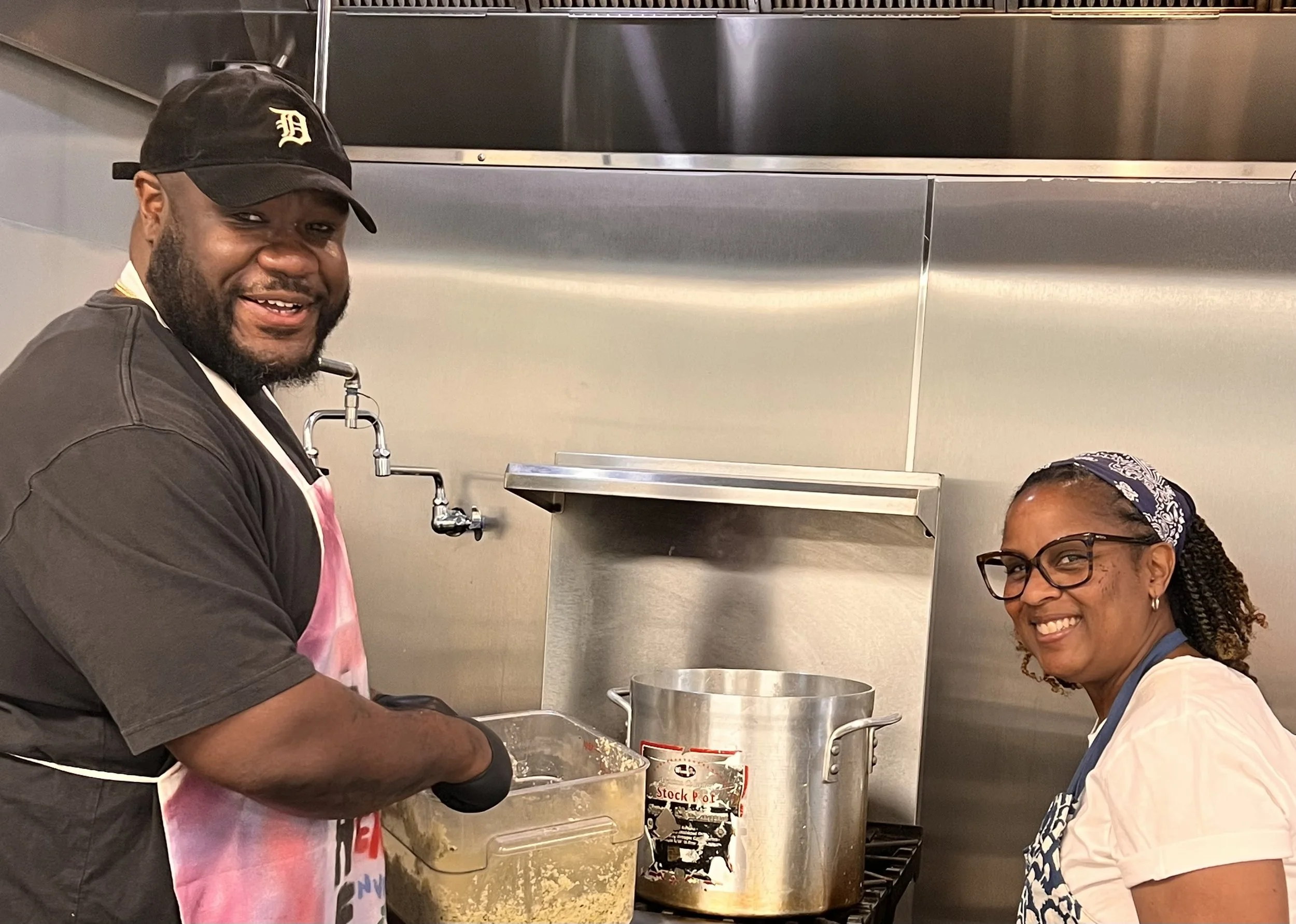 A man and woman smiling in a commercial kitchen, working with large cooking pots and containers of food.
