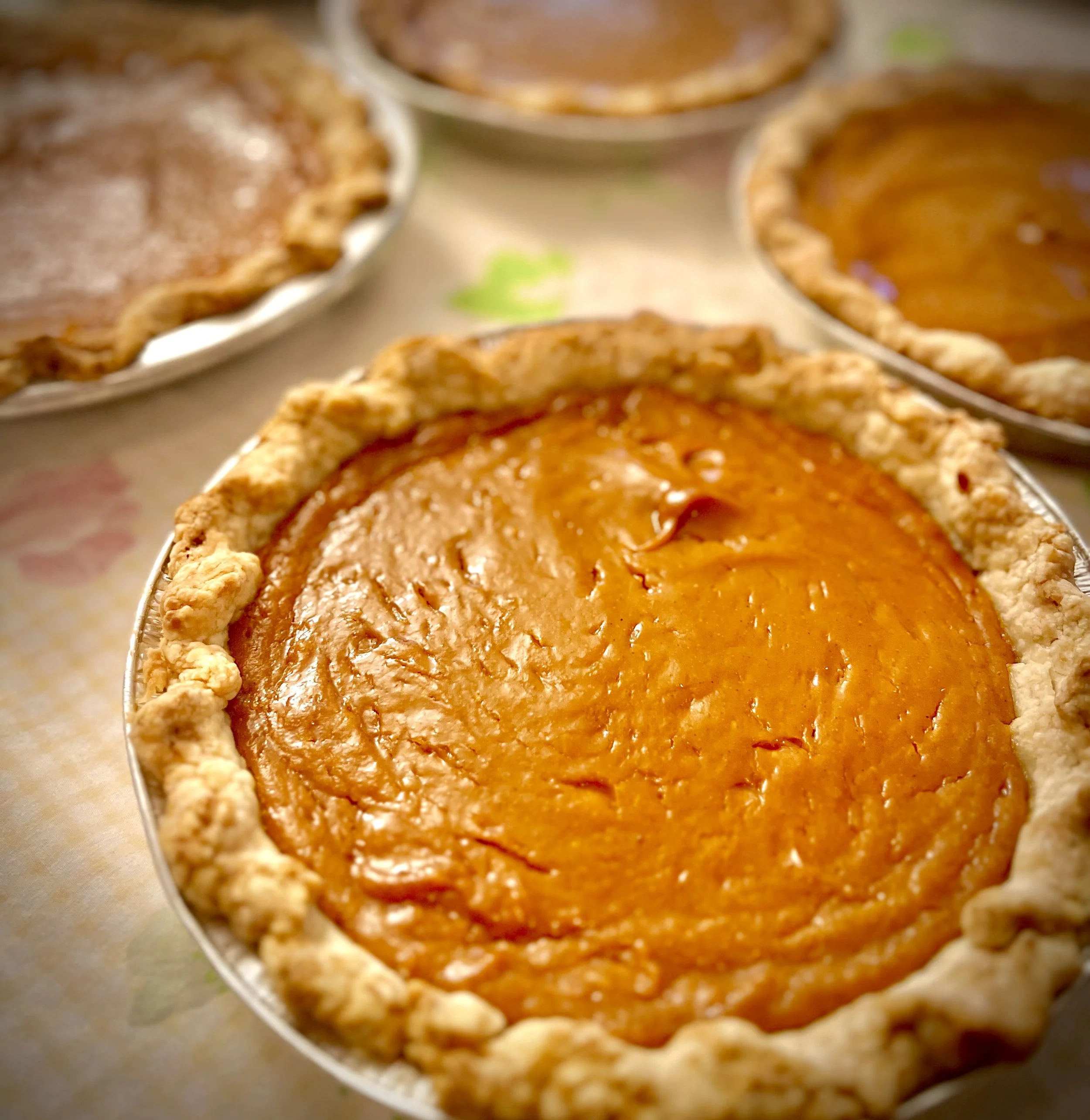 Close-up of pumpkin pie with a golden crust in a pie dish, with other pies in the background.