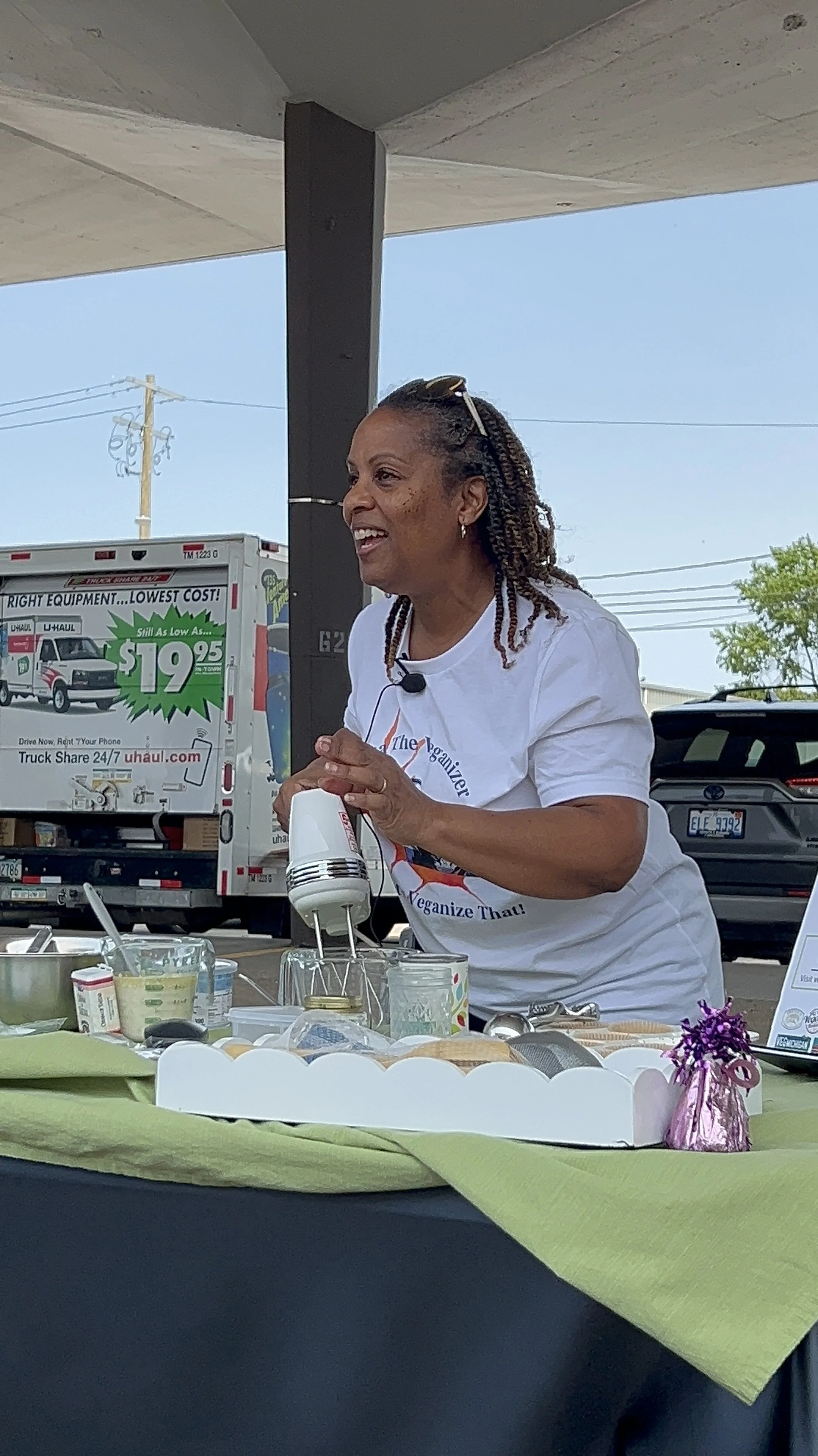 Woman demonstrating a cooking or baking process outdoors, smiling, using a hand mixer, with various ingredients and utensils on a table covered with a green cloth.