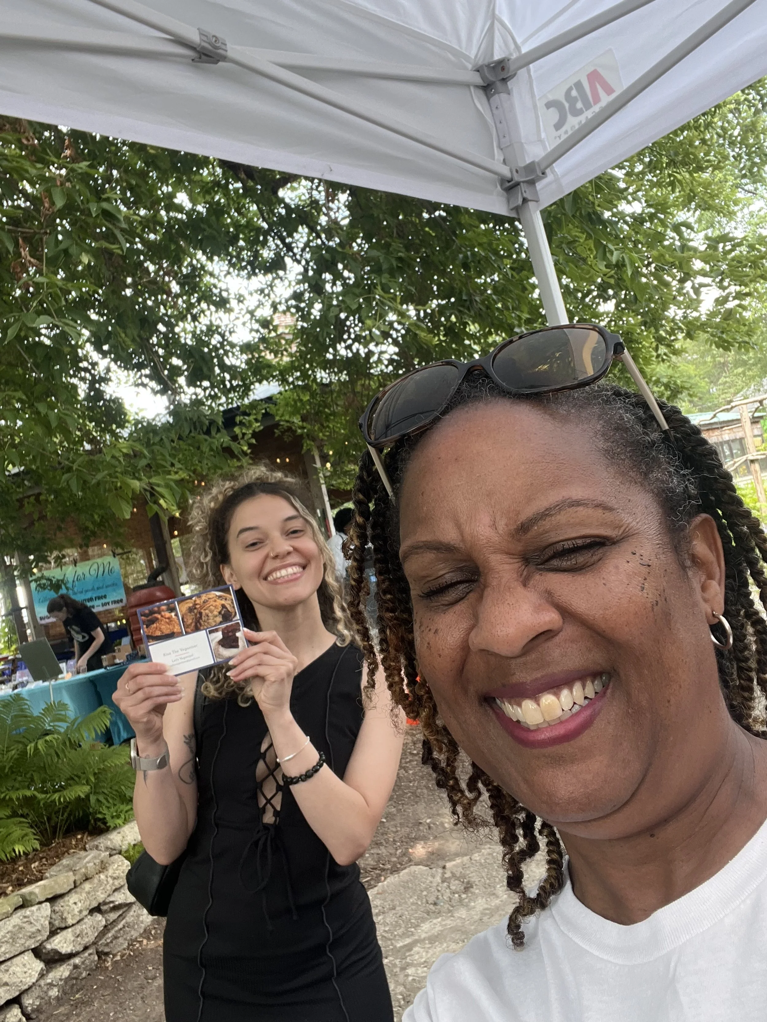 Two women smiling outdoors beneath a white canopy. The woman in the foreground has glasses on her head, dark curly hair, and is wearing earrings and a white shirt. The woman in the background has curly hair, is wearing a black sleeveless dress, and i