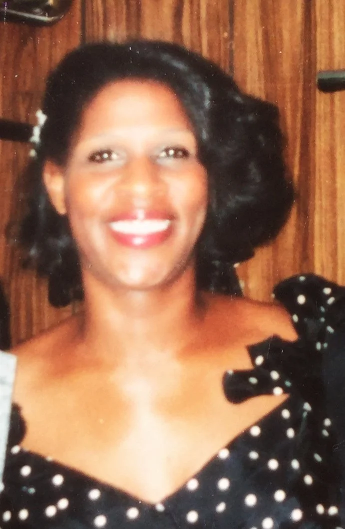 A woman with medium skin tone, black hair styled in waves, smiling and wearing a black dress with white polka dots, in front of a wooden background.