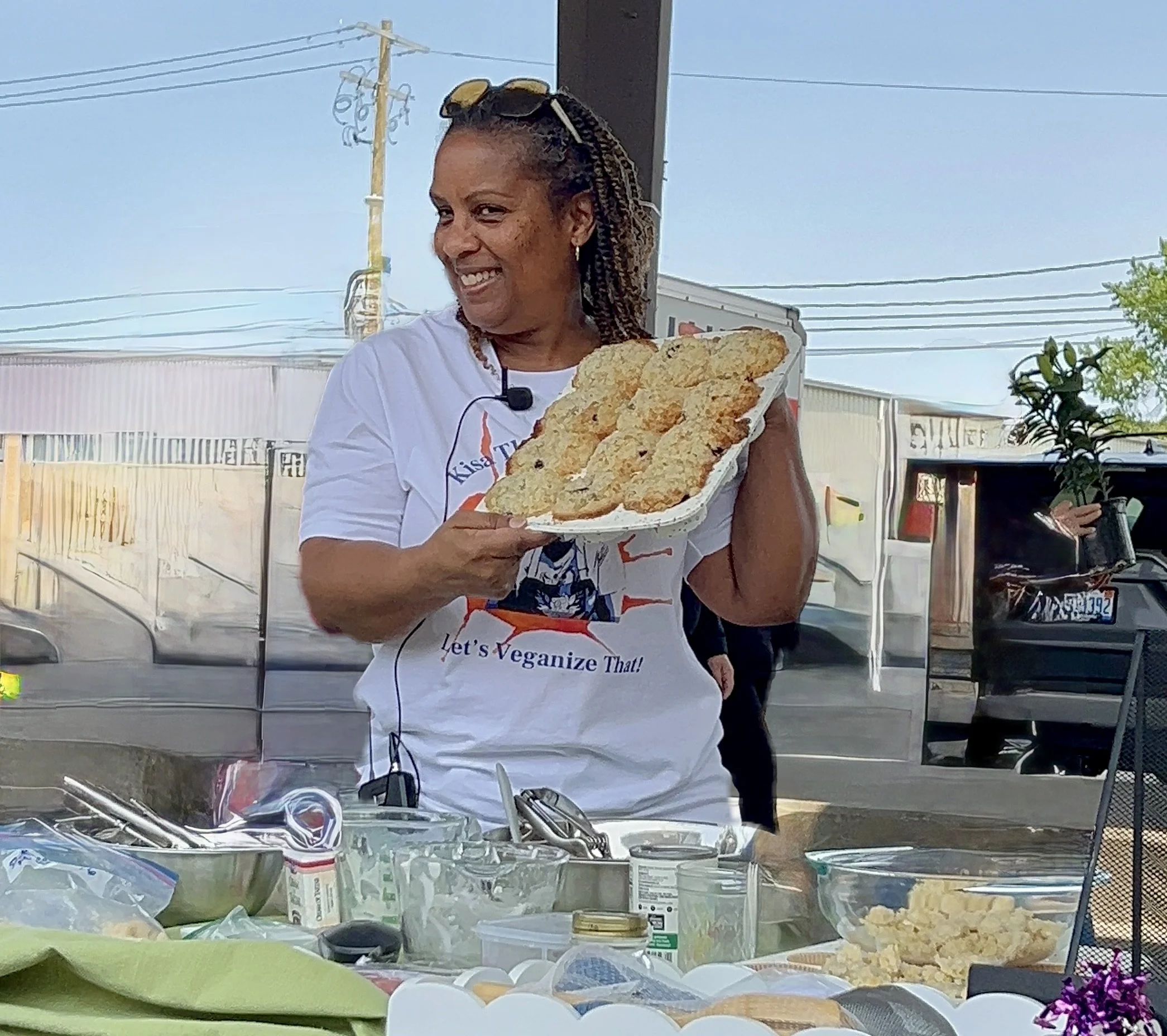 A woman smiling and holding a tray of breaded, fried food, standing behind a table with various kitchen utensils and ingredients, outdoors in a parking lot.