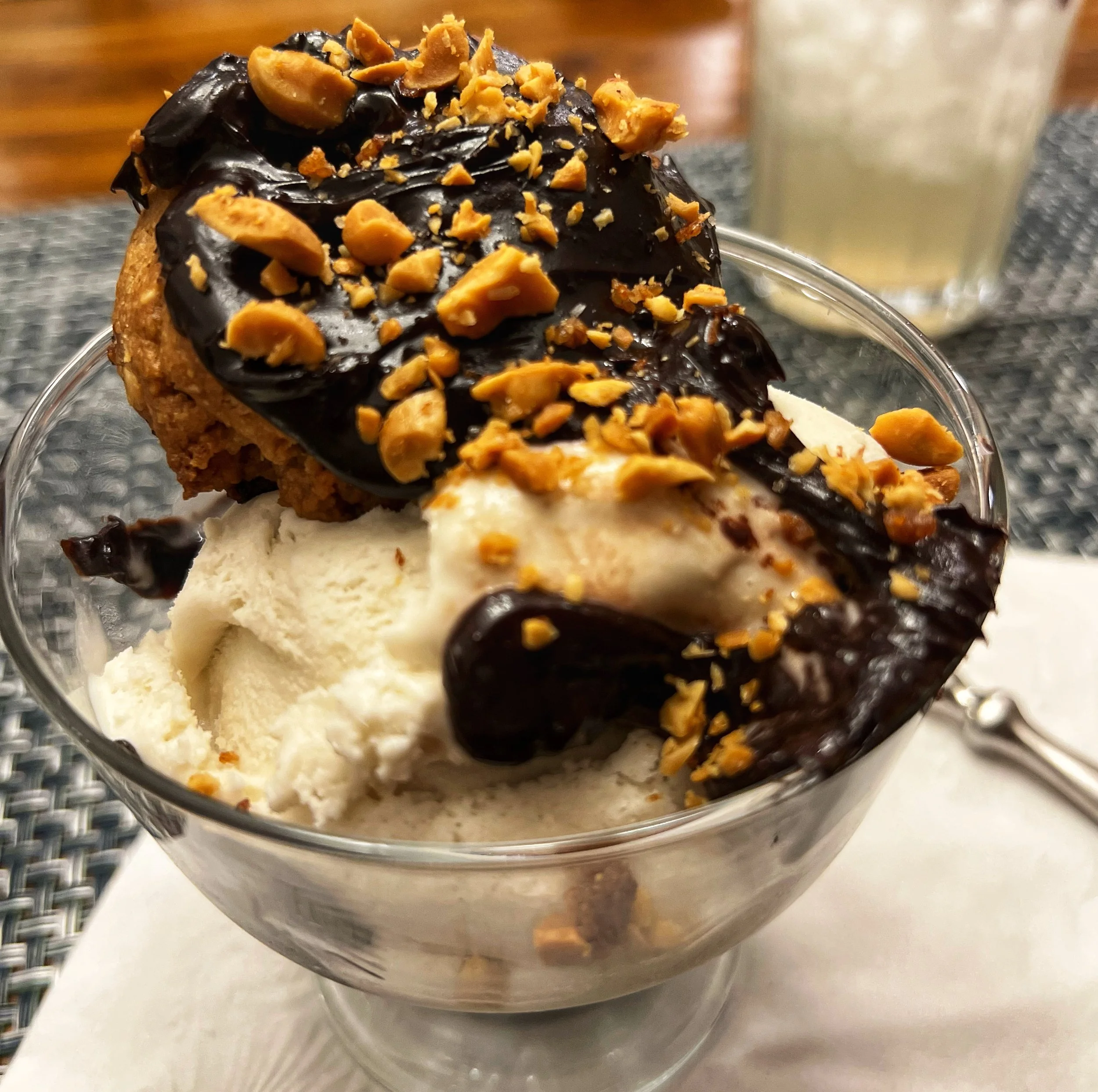 A dessert sundae in a clear glass bowl with vanilla ice cream topped with a large chocolate chip cookie, chocolate sauce, chopped peanuts, and a rock of chocolate ice cream in the background.