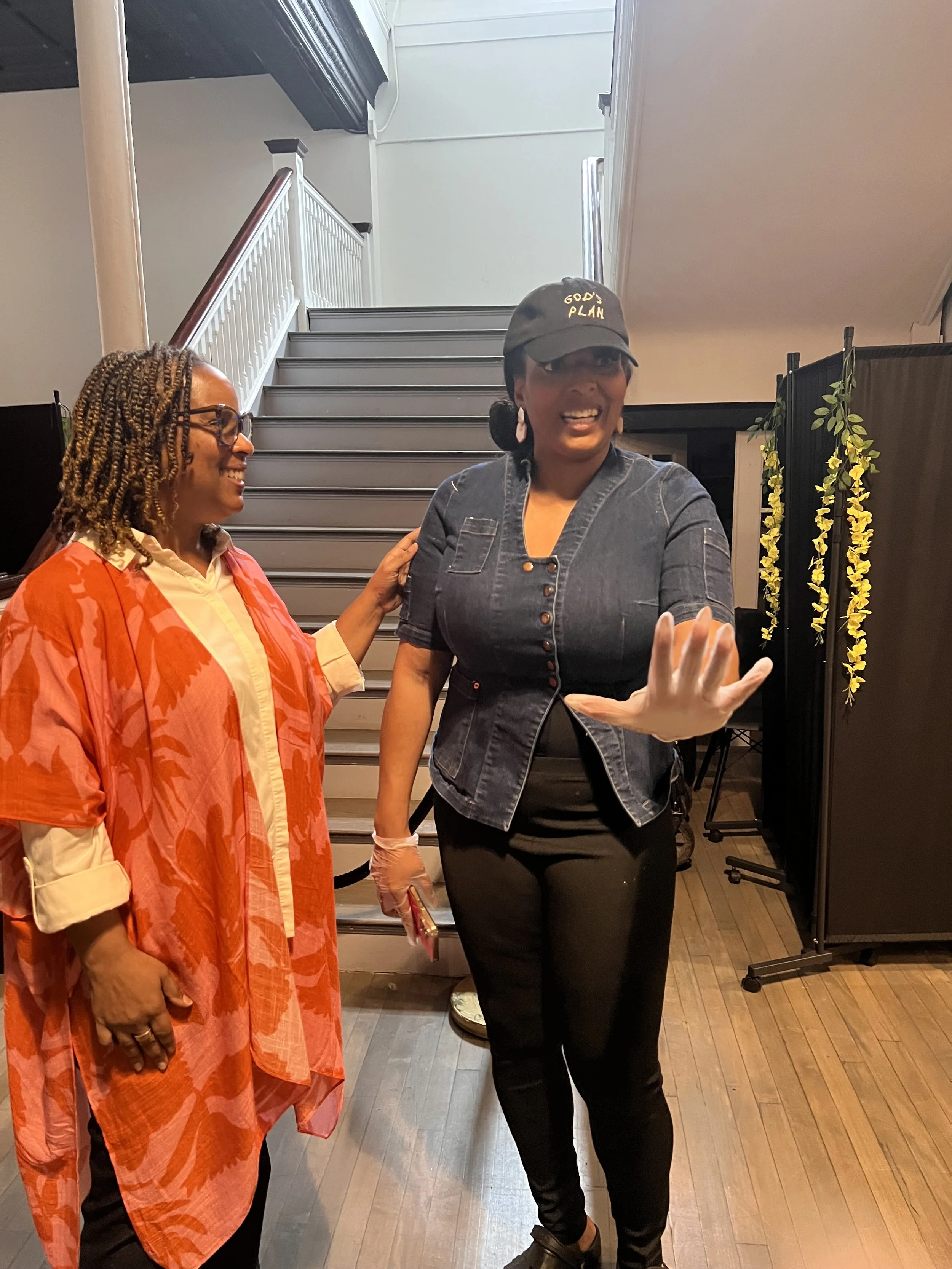 Two women smiling and talking indoors near staircase, one wearing a red patterned jacket, the other in a black cap with 'God's Plan' and a denim jacket.