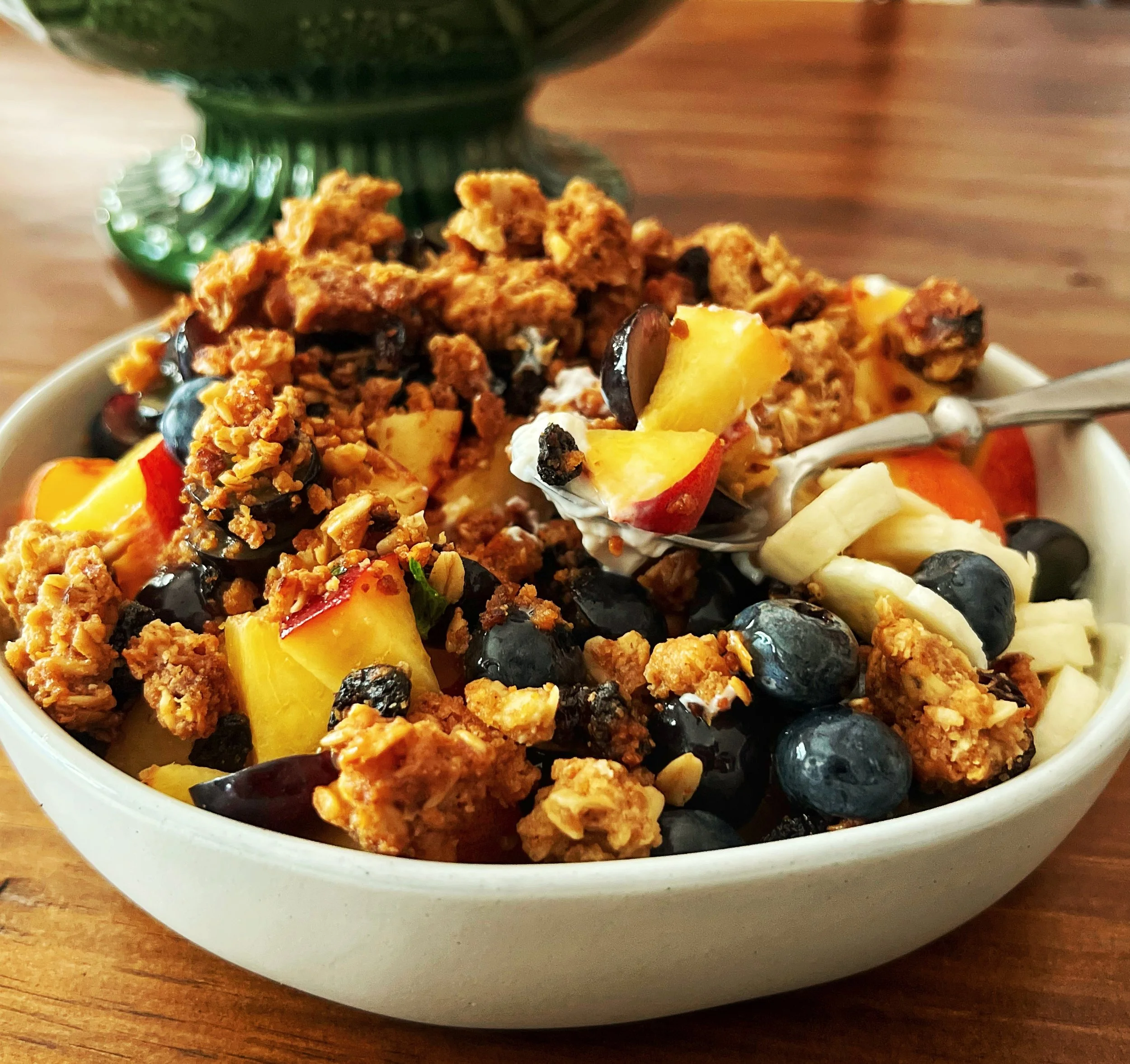 A bowl of granola with blueberries, chopped peaches, bananas, and granola with a spoon in a white bowl on a wooden table, with a green decorative item in the background.