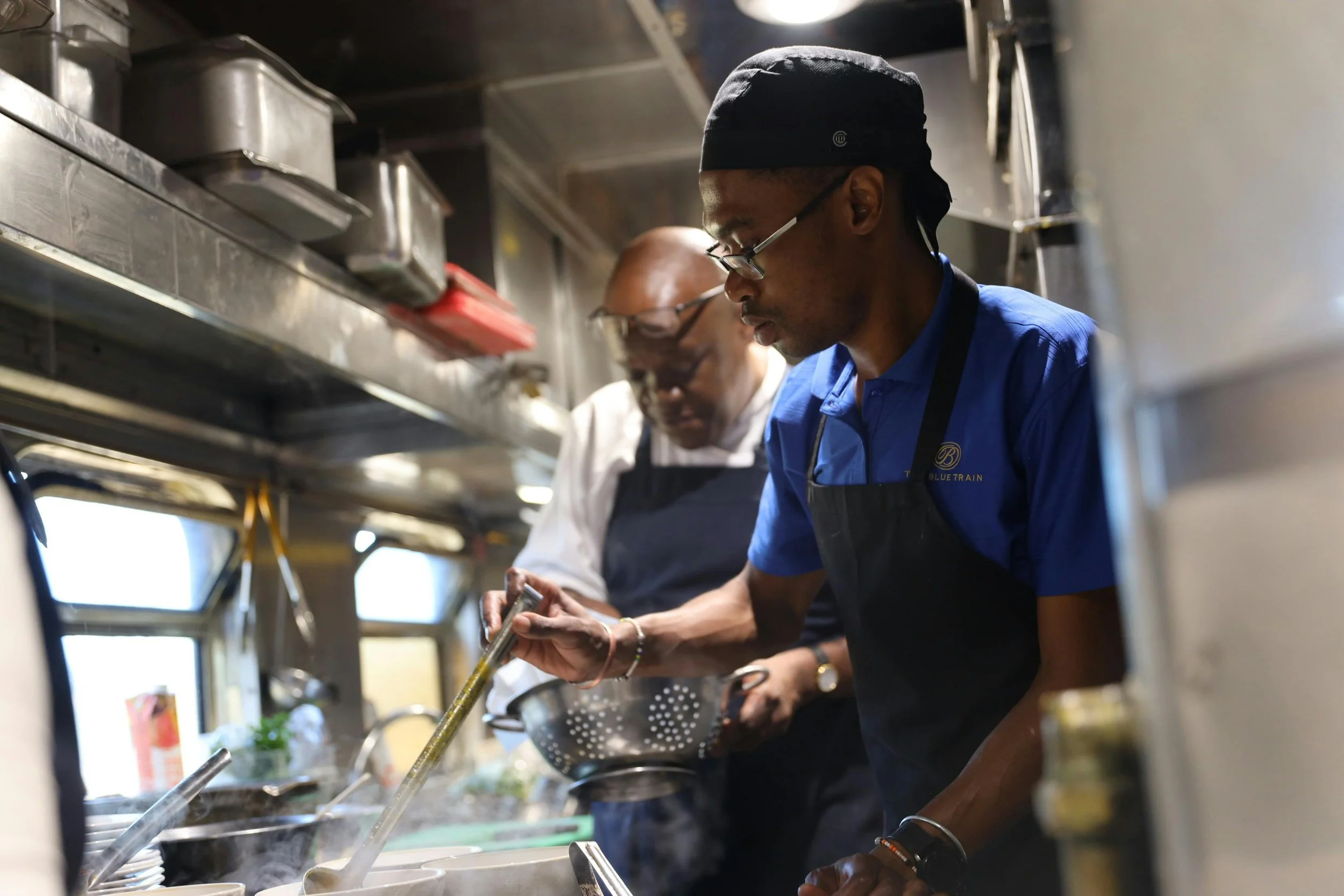 Two chefs cooking in a commercial kitchen, one stirring a pot and the other holding a colander.