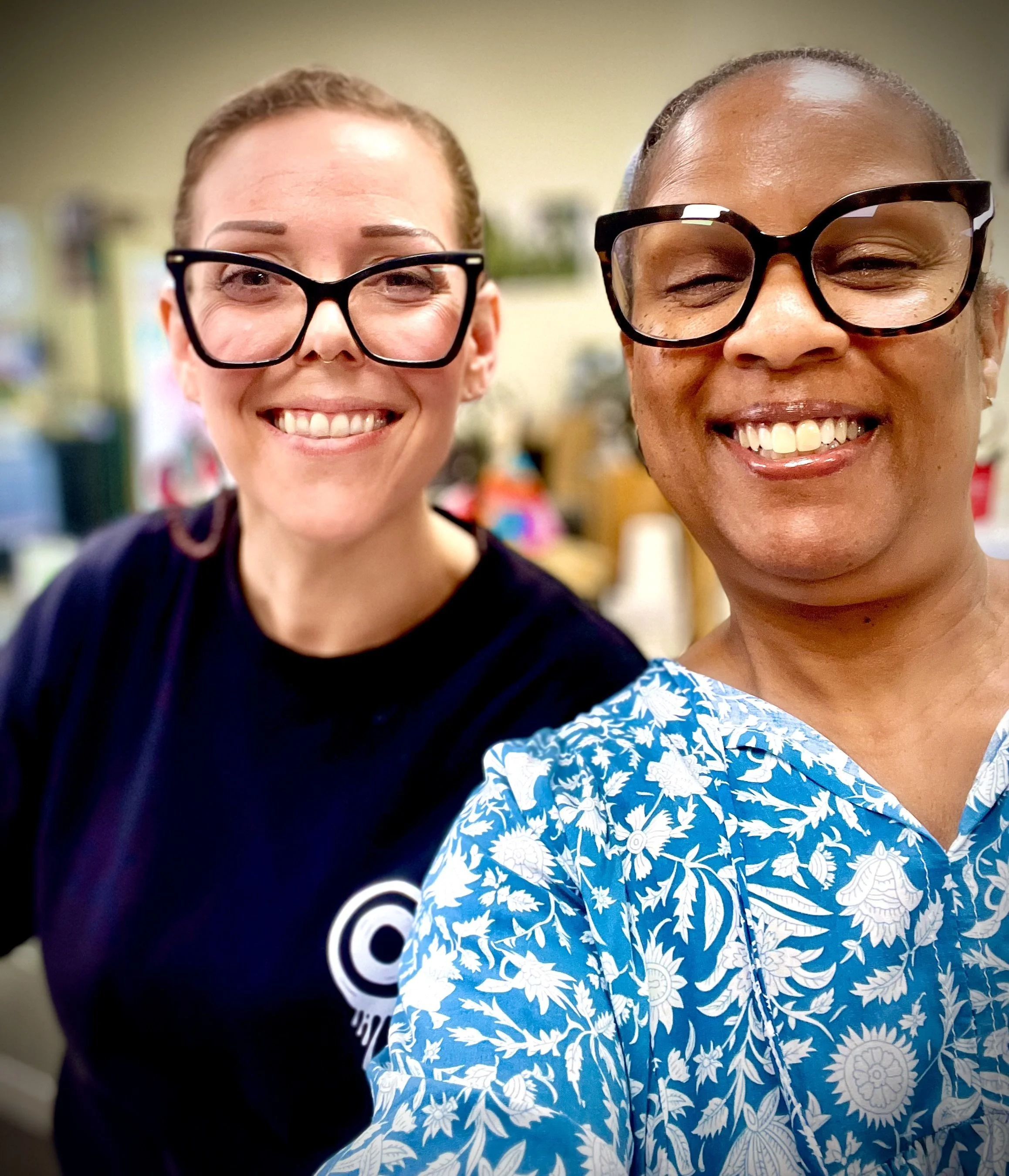 Two women smiling for a selfie, both wearing glasses, one with a black shirt and the other with a blue and white patterned shirt, in an indoor setting.