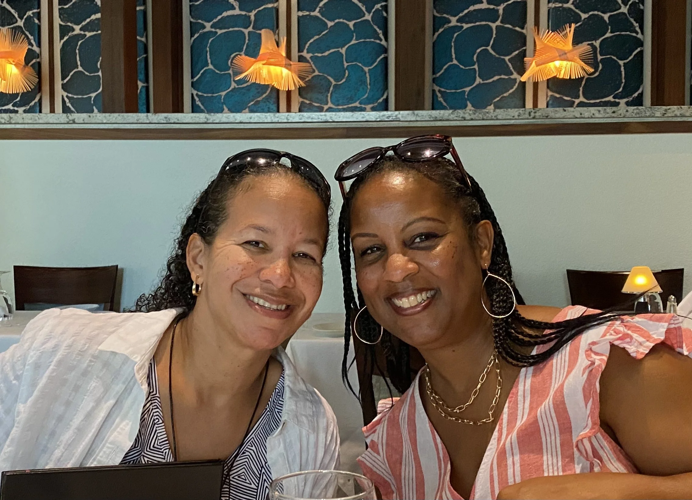 Two women smiling, sitting close together at a dining table in a restaurant, with glasses and tableware visible.