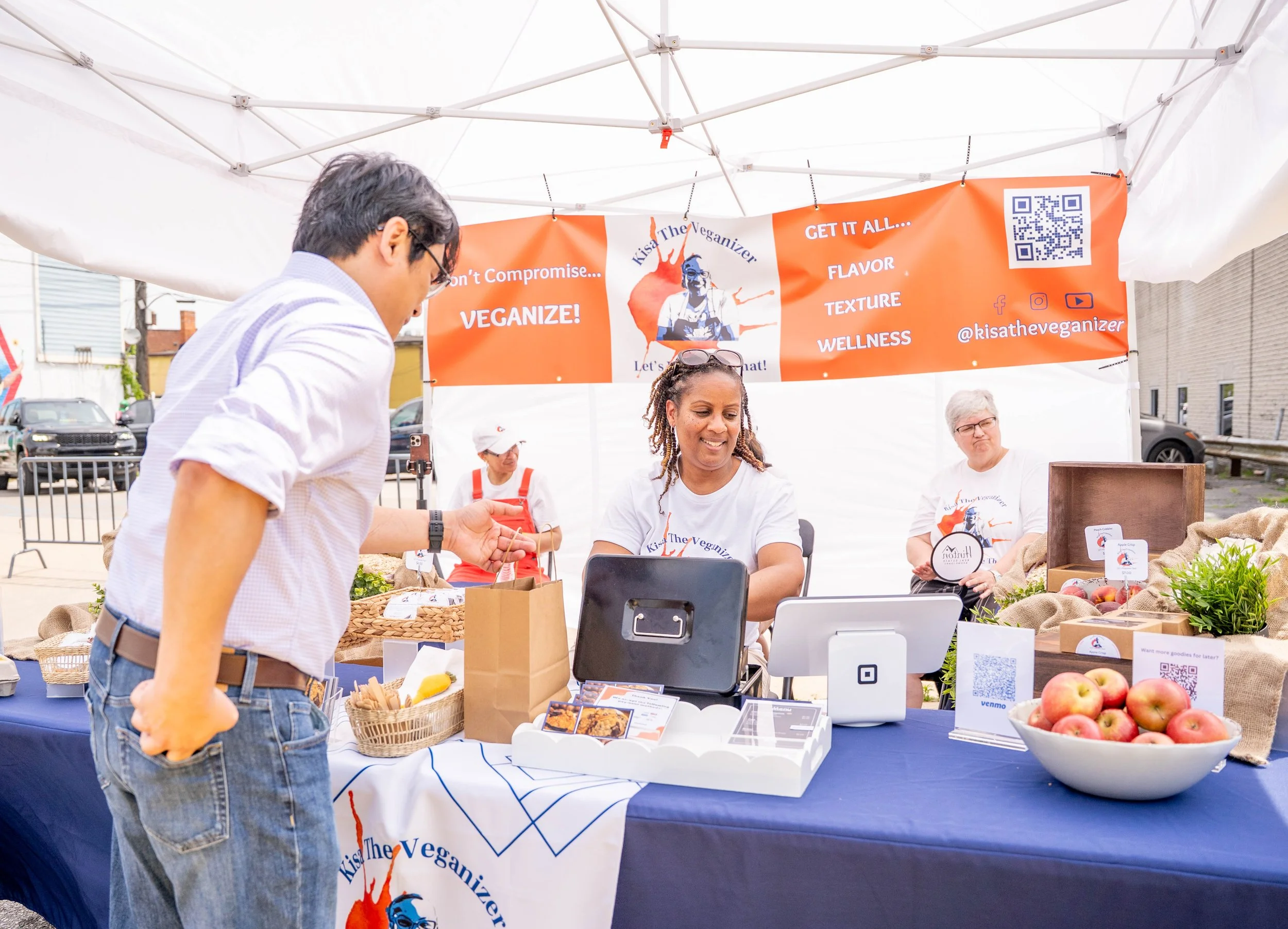 A man purchasing items from a vendor at a vegan market booth. The booth is under a white tent with vibrant orange signage that reads, 'Kiss The Veganizer,' and promotes vegan options with phrases like 'Don't Compromise... VEGANIZE!' and 'GET IT ALL..