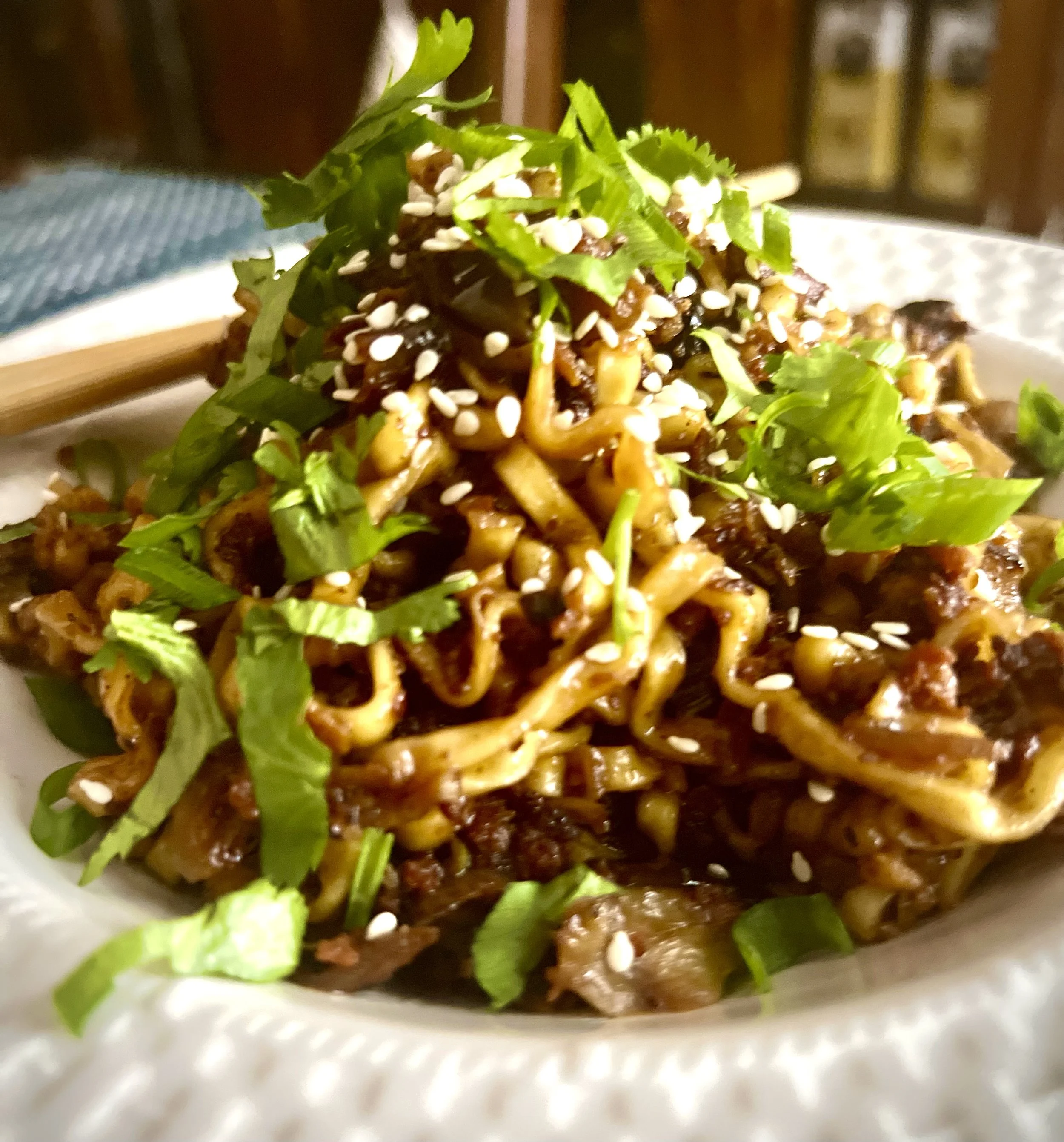Close-up of a bowl of Asian noodles topped with green herbs, sesame seeds, and a dark sauce.
