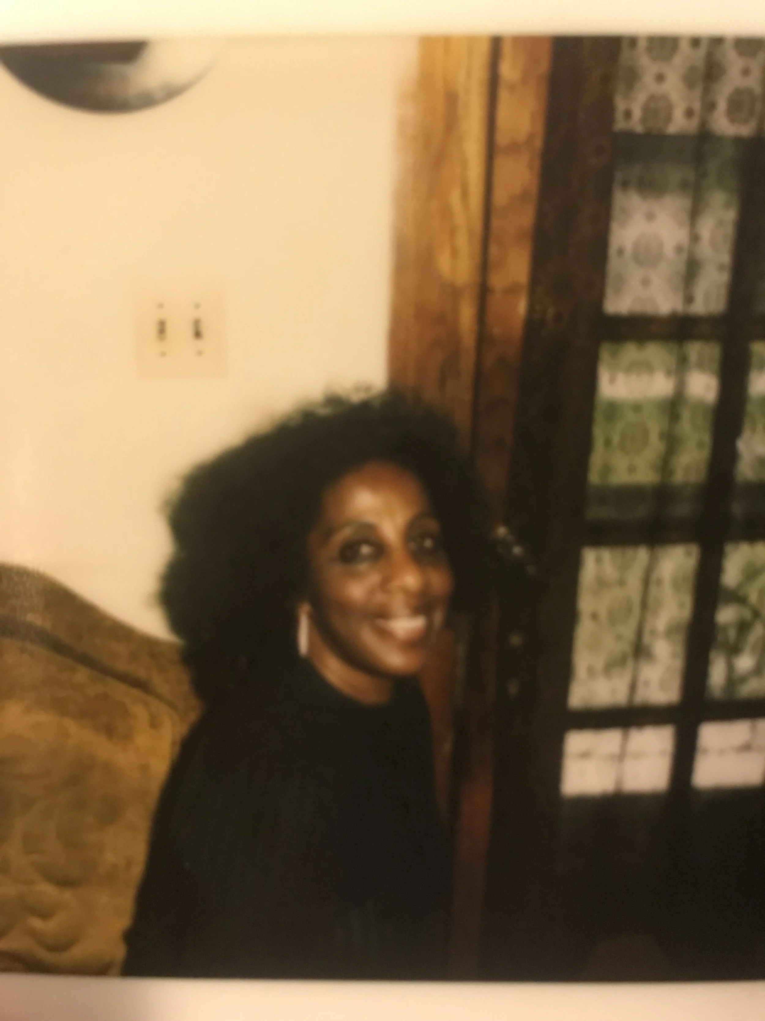 A woman with dark, curly hair, wearing earrings and a dark top, smiling while sitting on a sofa in a room with a wooden door and patterned curtains in the background.