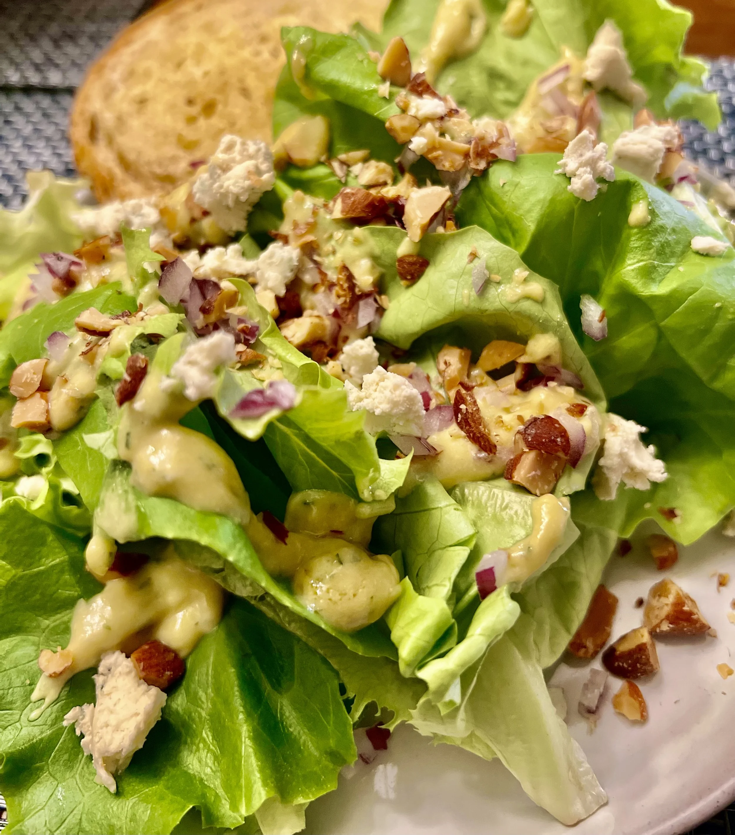 Close-up of a fresh salad with lettuce, chopped nuts, crumbled cheese, and a creamy dressing, served on a white plate with some bread in the background.
