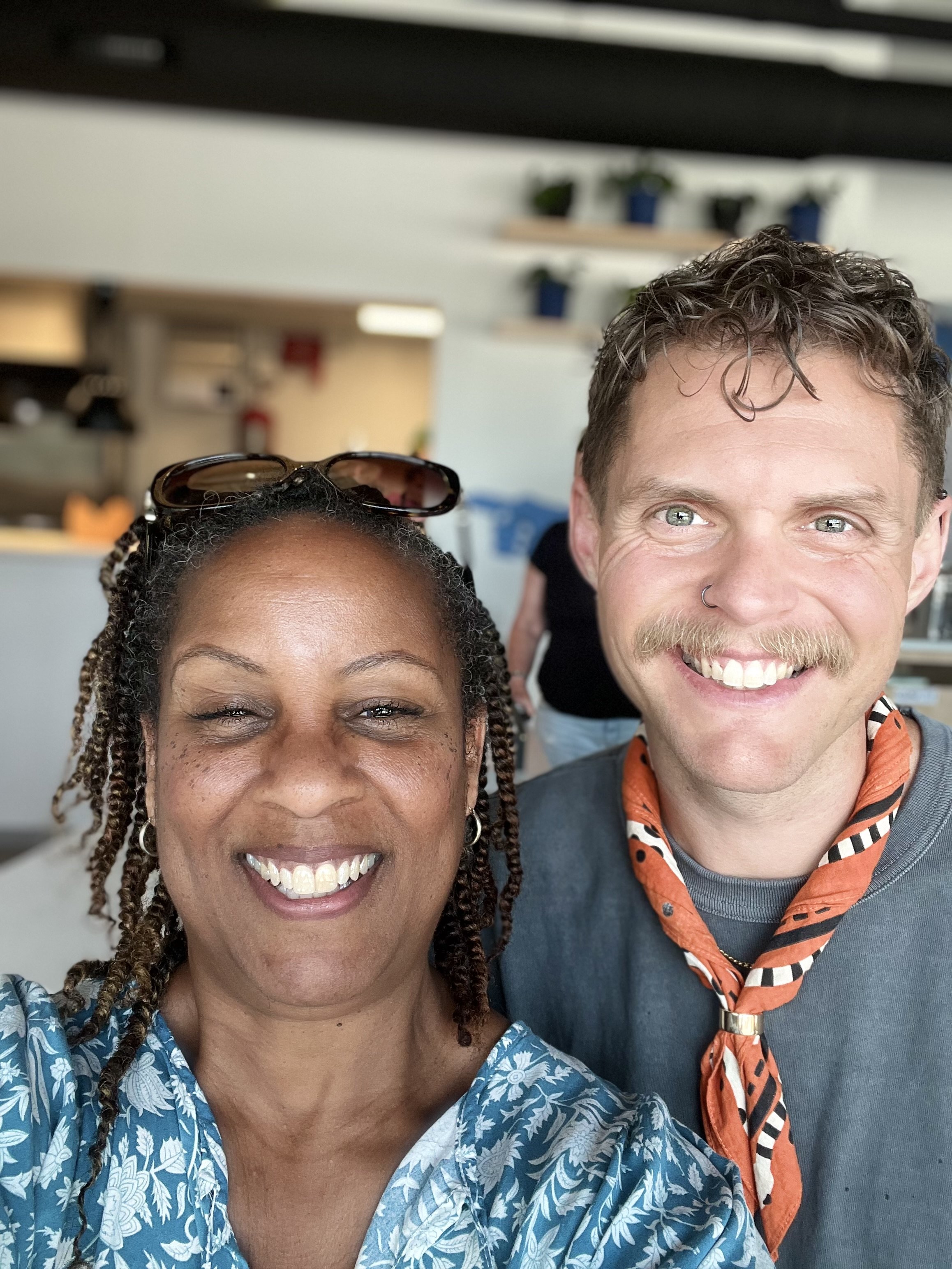 A woman and a man smiling for a selfie indoors, with the woman on the left and the man on the right. The woman has dark skin, braided hair, and sunglasses on her head. The man has light skin, curly hair, a mustache, a nose ring, and is wearing a gray