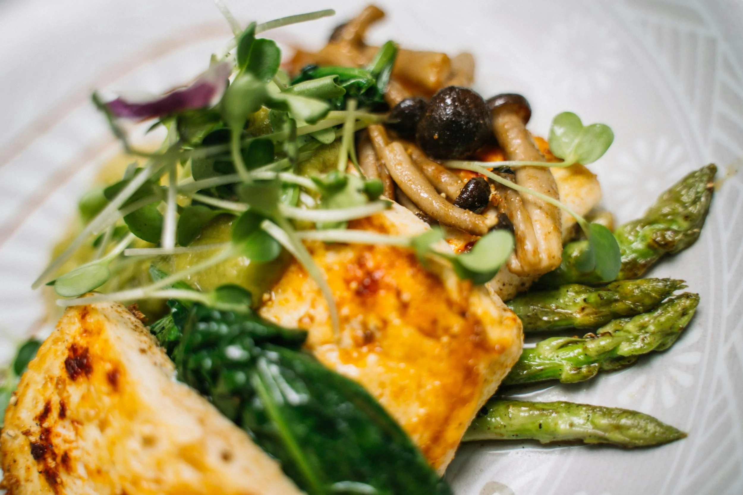 Close-up of a plate of grilled chicken with herbs, asparagus, sautéed mushrooms, microgreens, and avocado slices.