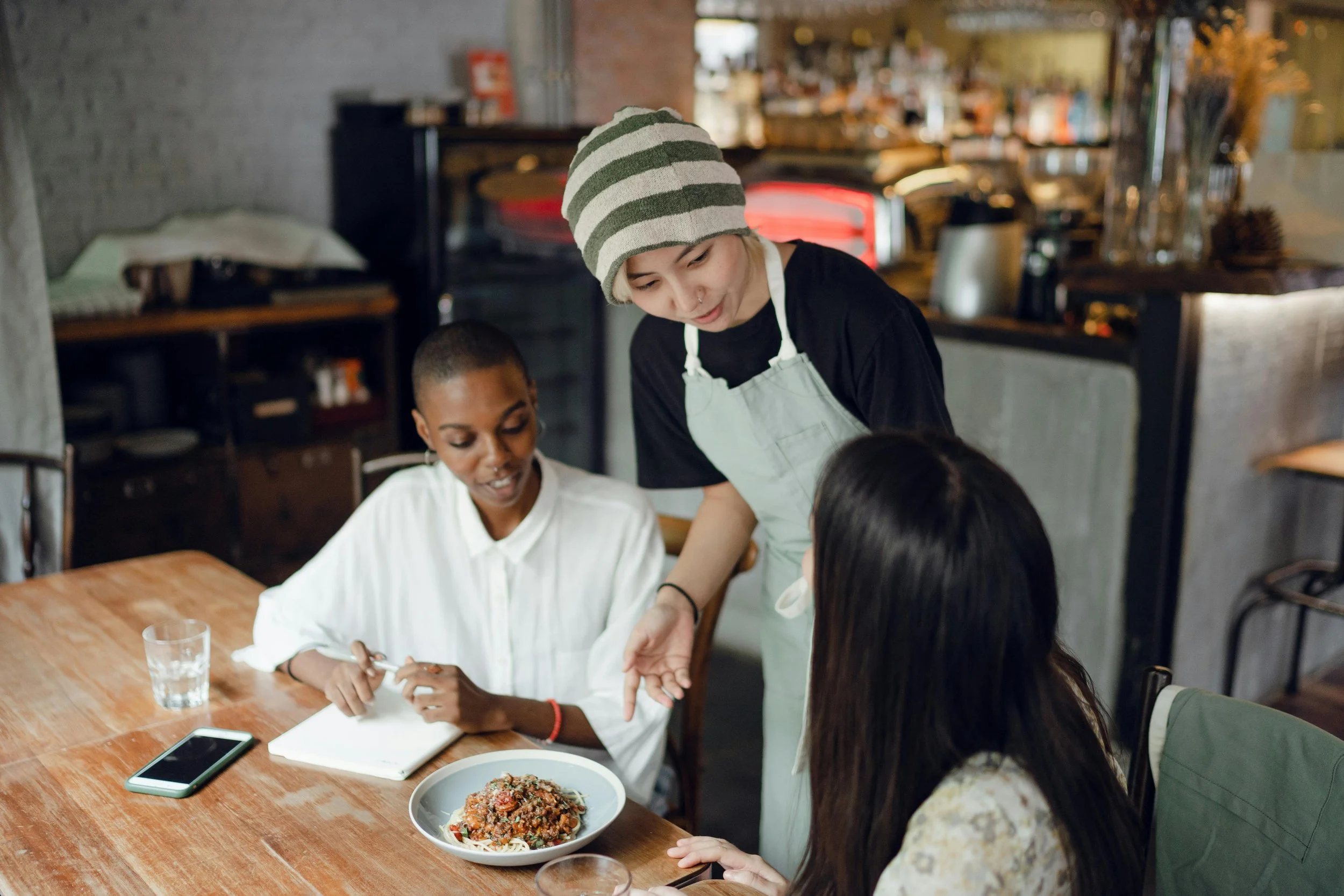 A woman with a striped beanie and white apron showing a plate of food to two women at a wooden table in a cozy restaurant.