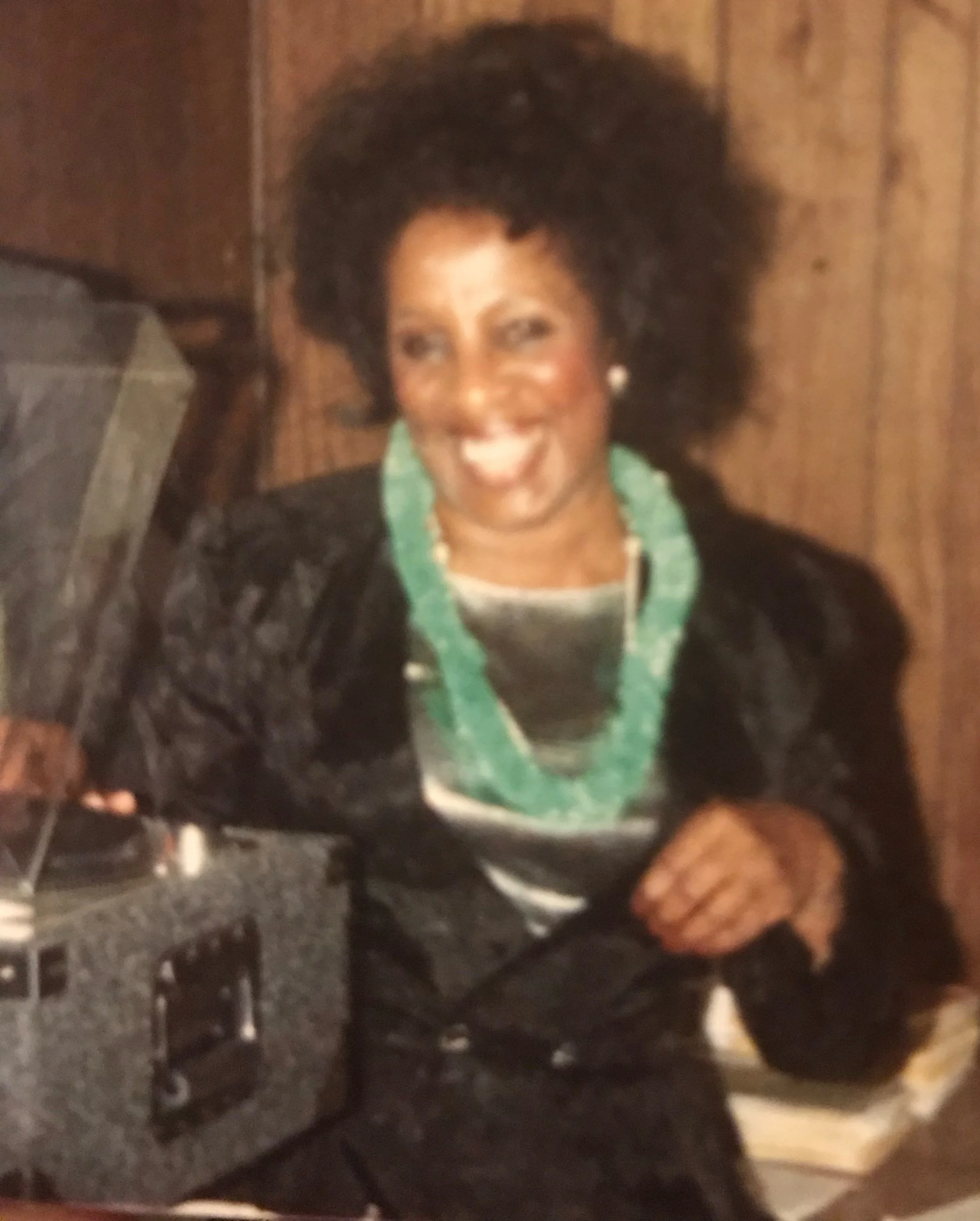 A smiling woman with dark curly hair, wearing a black jacket and green necklace, sitting at a table with a gramophone in front of her.