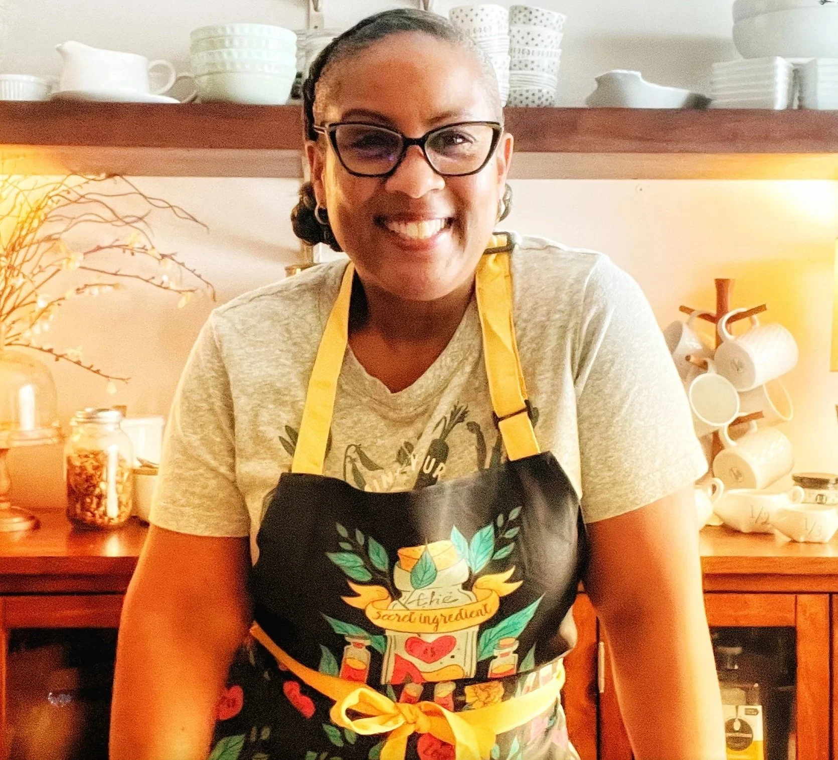 Smiling woman wearing glasses and a gray T-shirt, standing in a kitchen with a wooden cabinet, white dishes, and cups in the background, wearing a black apron with colorful design and yellow ribbon