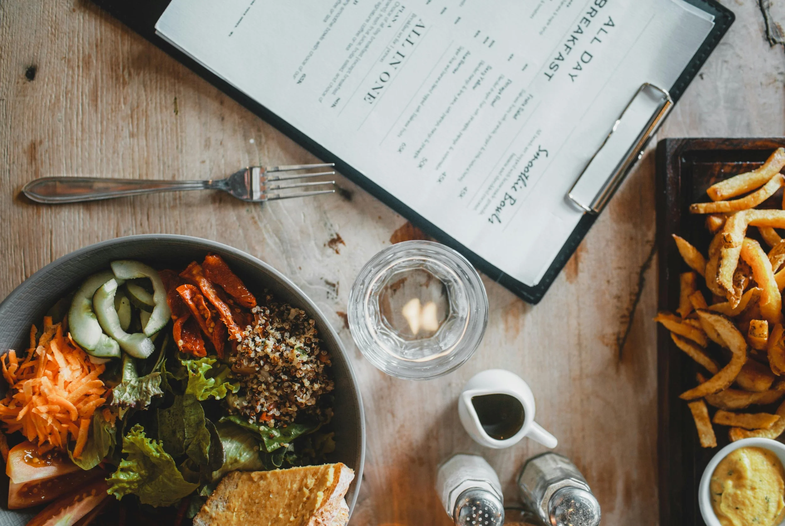 A restaurant table with a salad, fries, a glass of water, a bottle of sauce, and a menu on a wooden table.