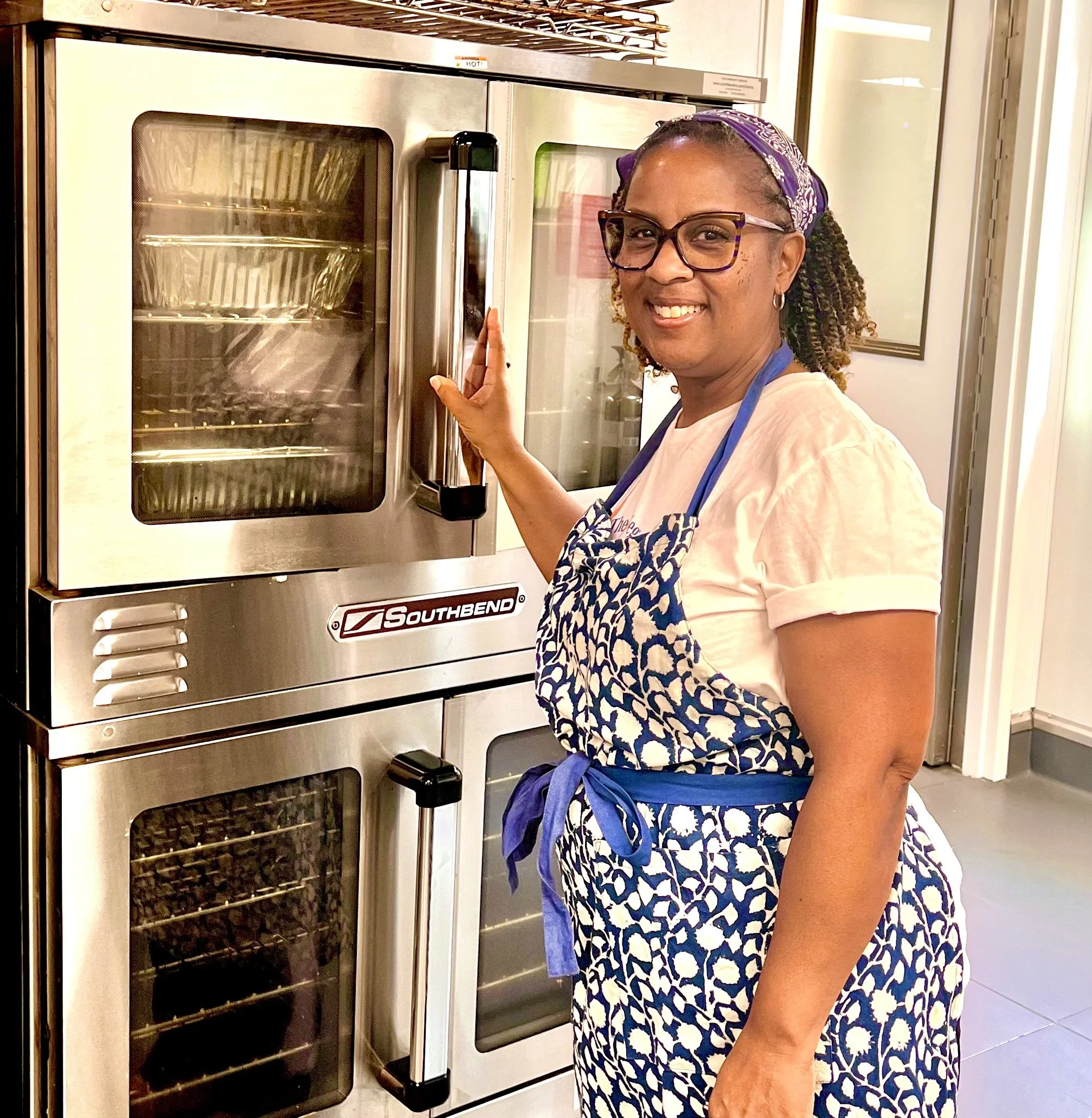 A woman wearing glasses and a purple headscarf, dressed in a white T-shirt and blue patterned apron, stands beside a commercial oven in a kitchen, smiling and holding the oven door handle.