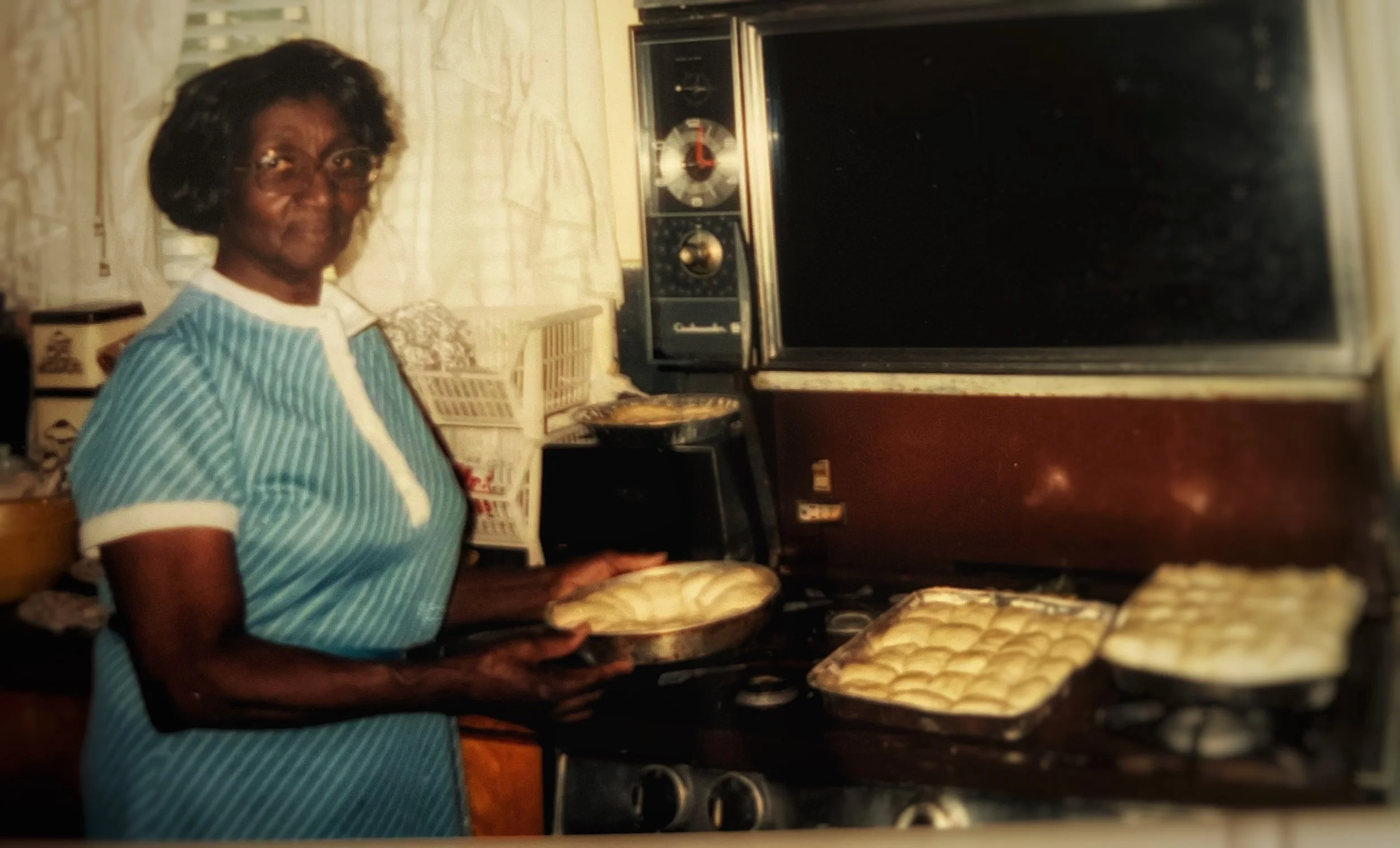 An elderly woman in a blue and white striped dress baking in a kitchen, holding a pie dish with a pie, with a baking tray of unbaked pastries on the stove, and a vintage TV and radio in the background.