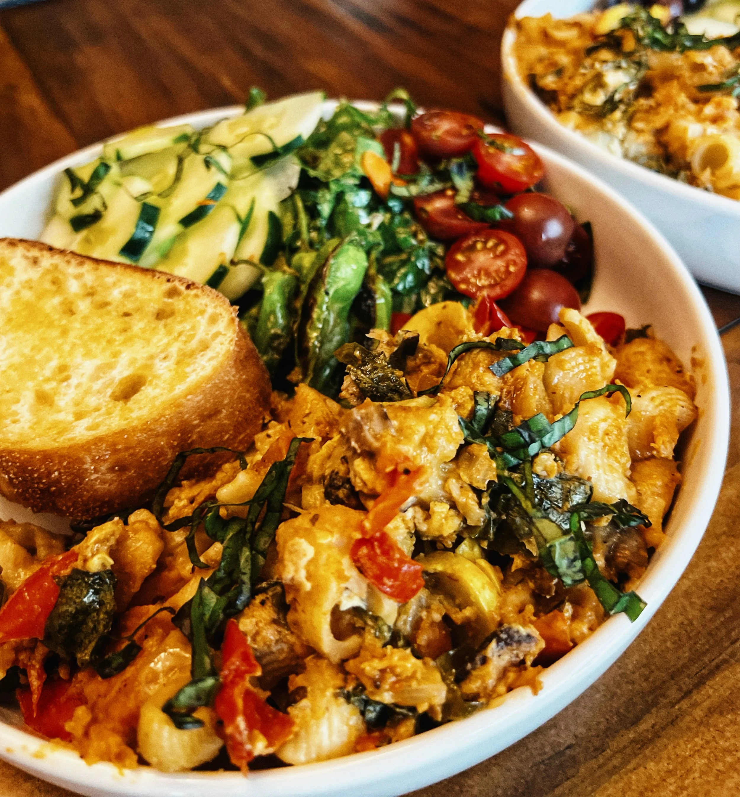 Mixed vegetable salad with cherry tomatoes, cucumber slices, shredded greens, a slice of bread, and a vegetable casserole in a white bowl.