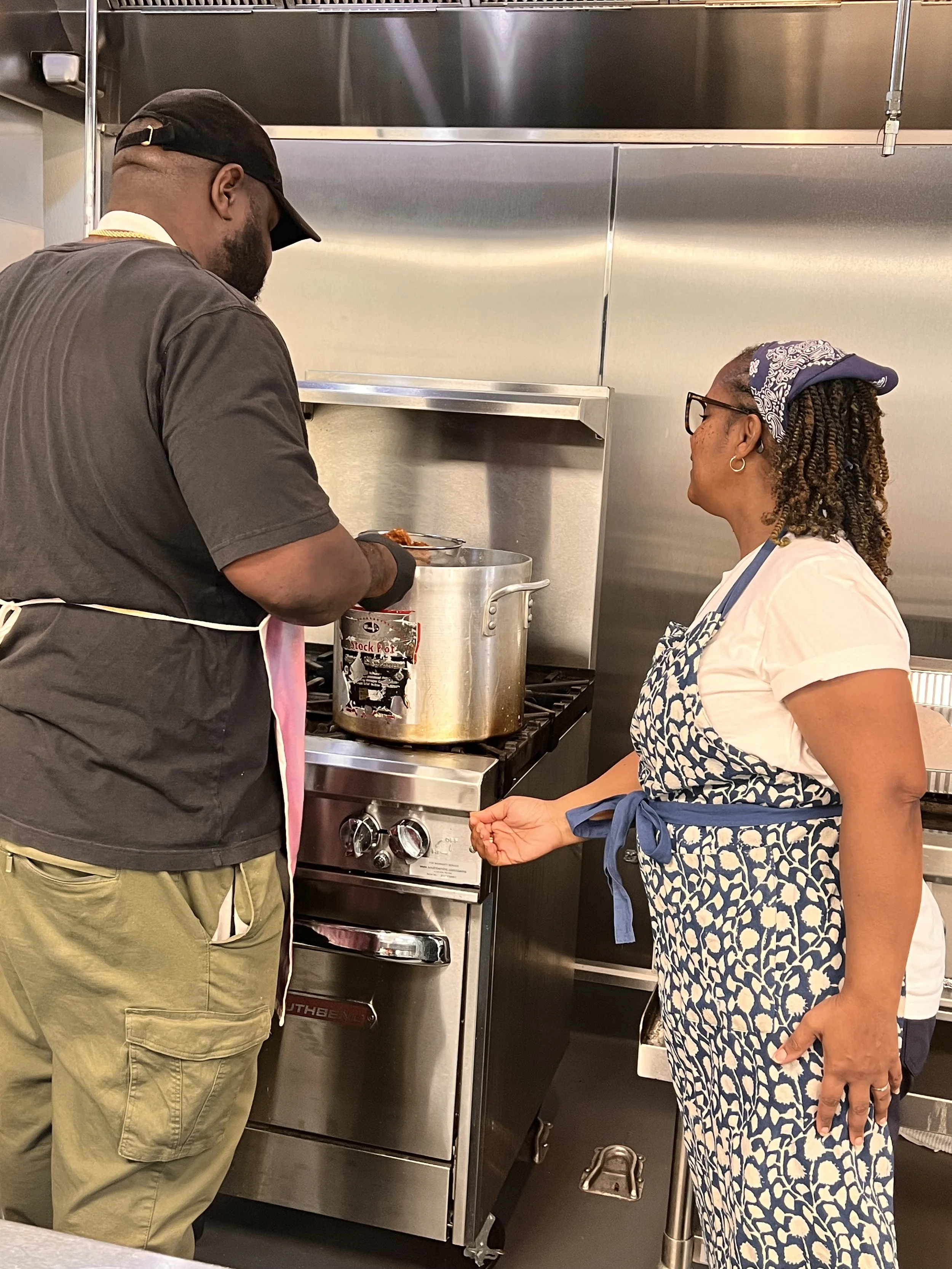 Two women in a kitchen, one is cooking with a large pot on the stove, and the other is standing and talking to her while wearing an apron and a headscarf.