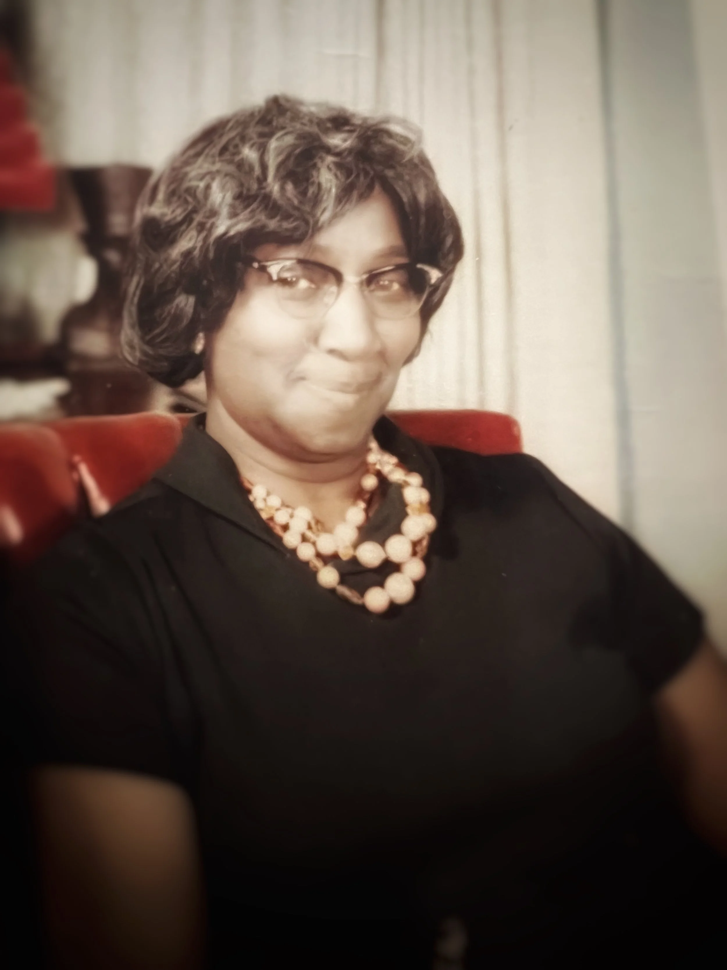A woman with short, curly hair, wearing glasses, a black top, and a pearl necklace, sitting on a red chair indoors.