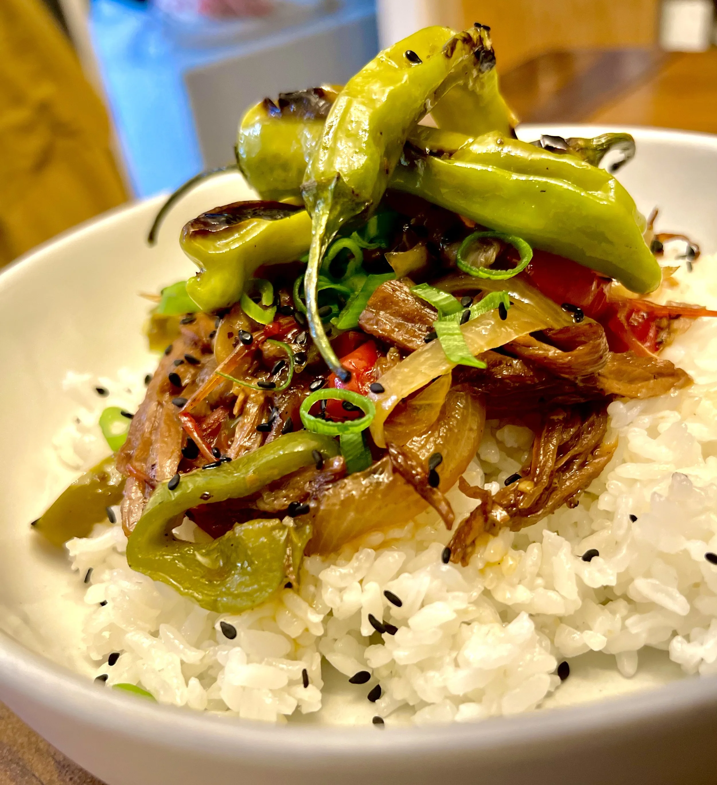 Bowl of white rice topped with grilled green peppers, shredded meat, chopped green onions, sesame seeds, and sauce.