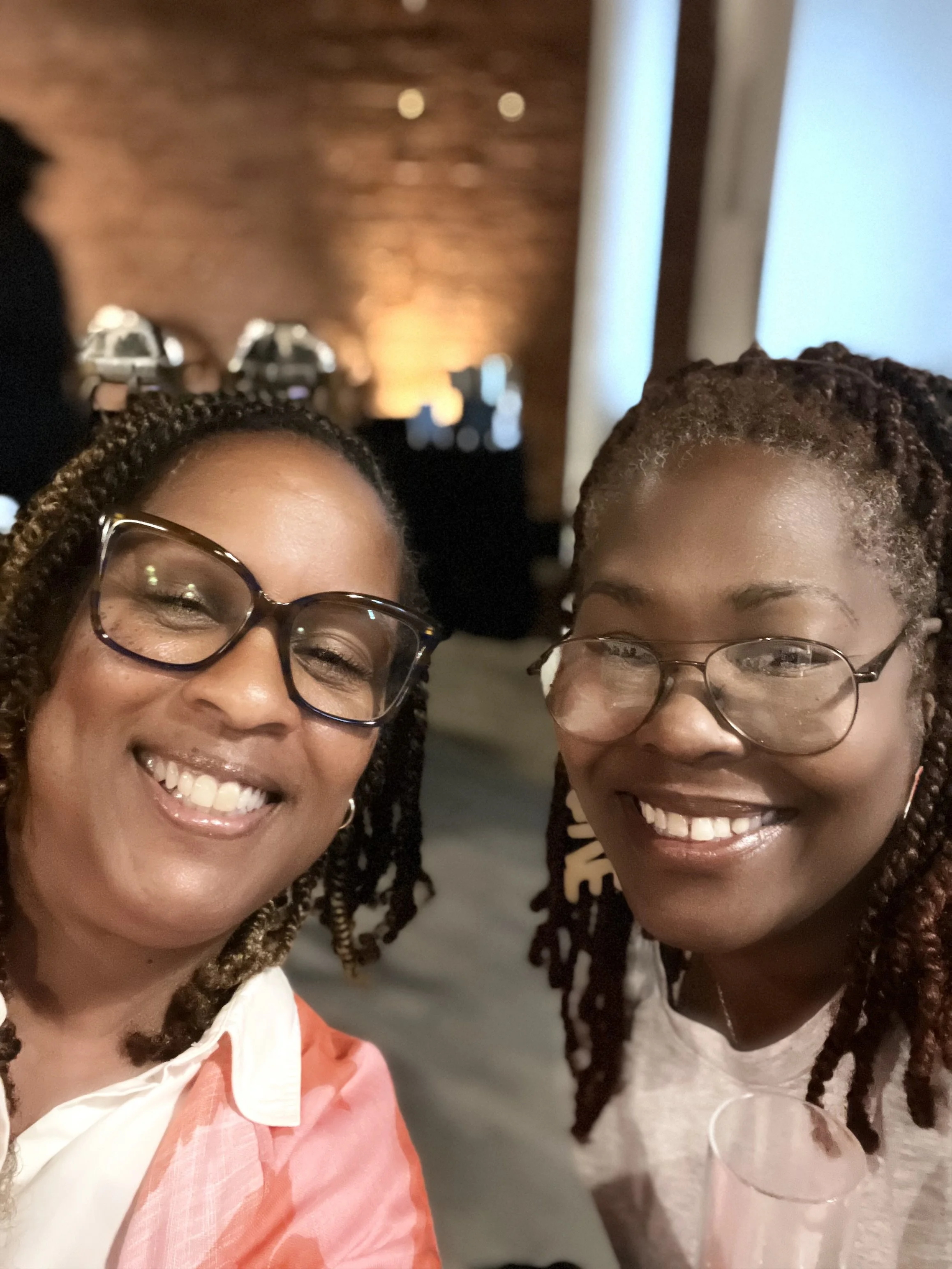 Two women smiling at a social event, wearing glasses, with a brick wall and blurred background behind them.