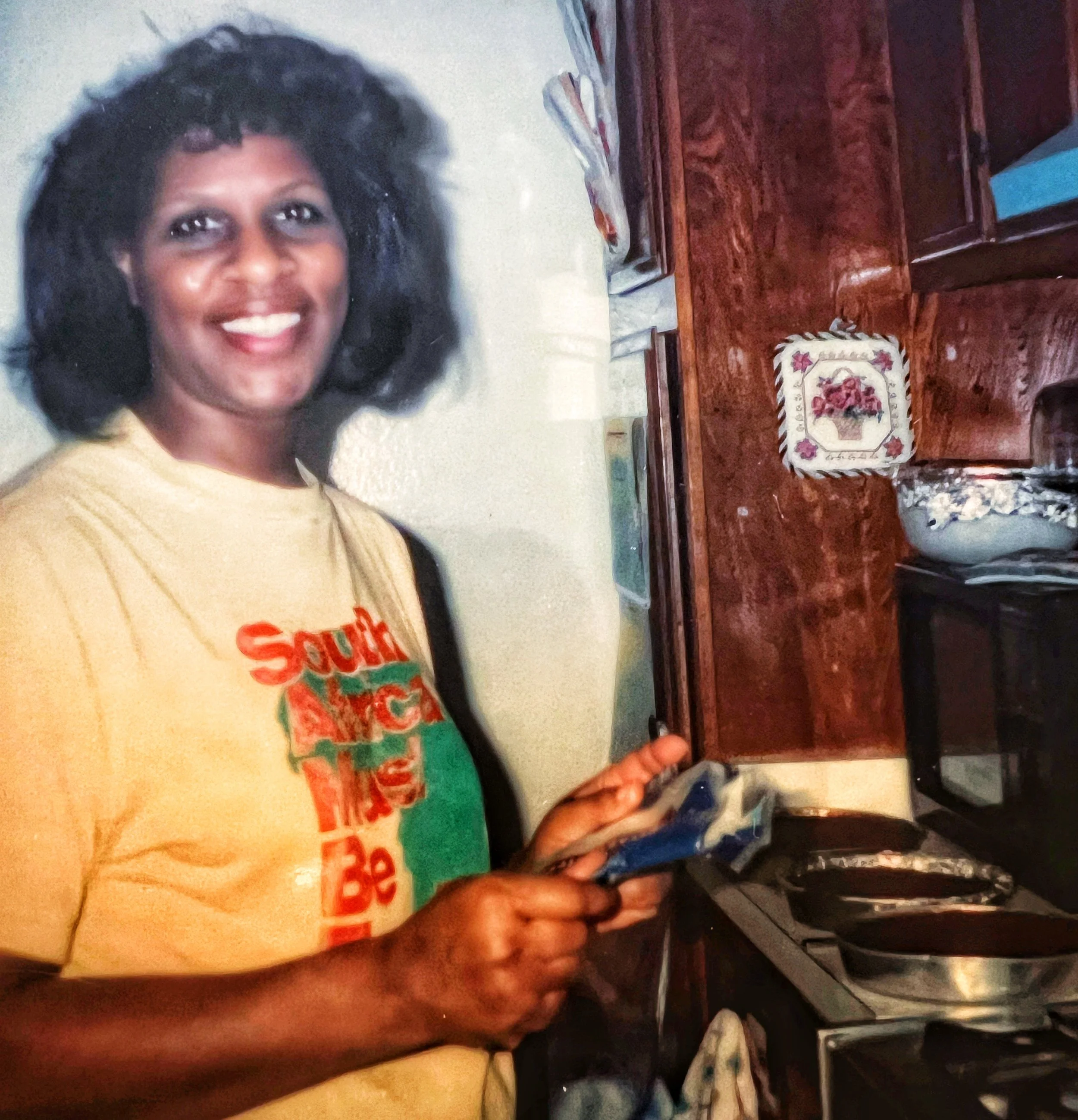 A woman with dark, curly hair smiling, wearing a yellow T-shirt with red and green graphics, standing in a kitchen with wooden cabinets, a small table with a decorative cloth, and a microwave oven.