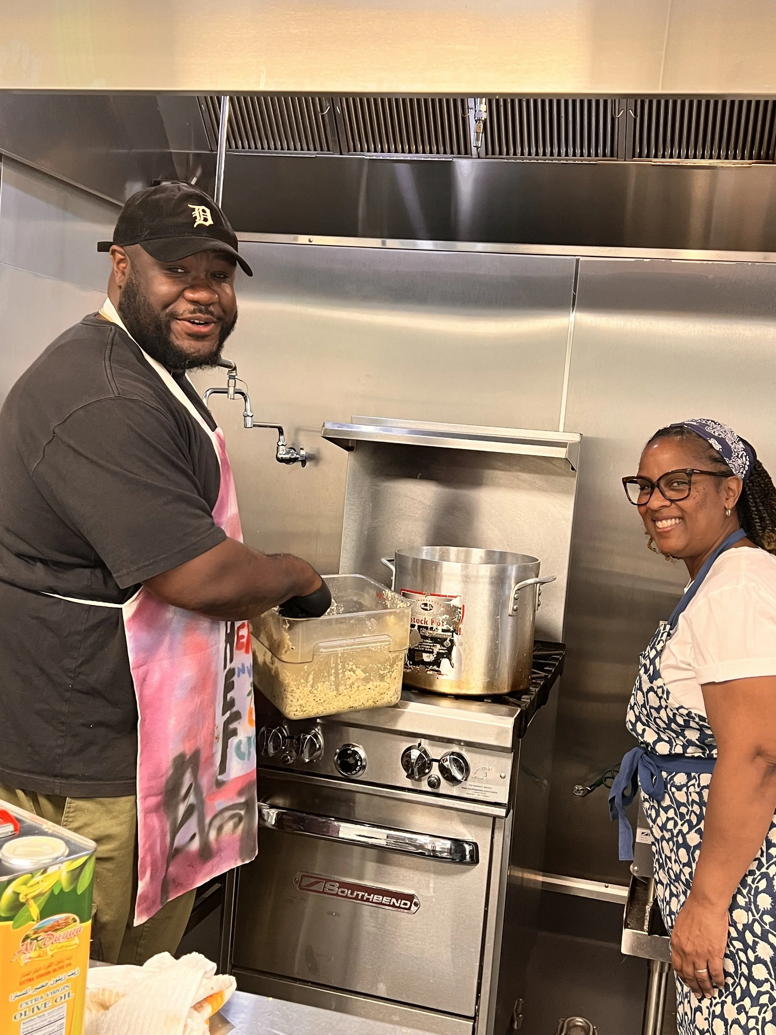 Two people cooking in a professional kitchen with stainless steel appliances. The man on the left wears a black cap, black shirt, and apron, and is stirring food in a clear container. The woman on the right wears glasses, a patterned apron, and a hea
