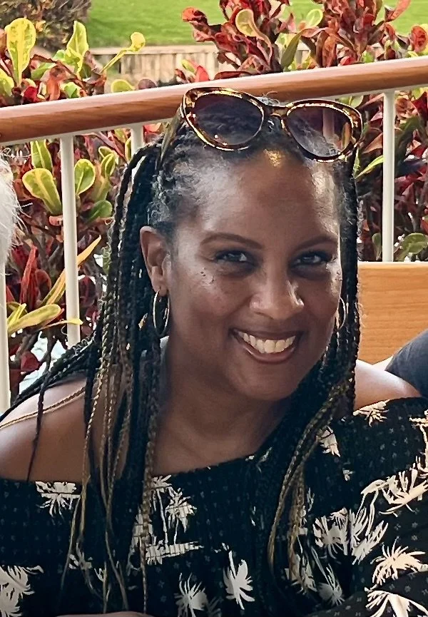 A woman with braided hair and sunglasses on her head, smiling outdoors near a railing with green and red plants in the background.