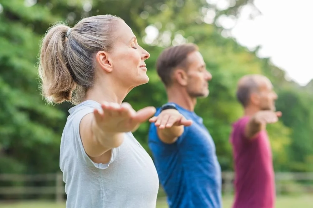 Tre persone adulte fanno esercizi di stretching all'aperto in un parco, con le braccia tese in avanti e occhi chiusi, sotto la luce del sole.