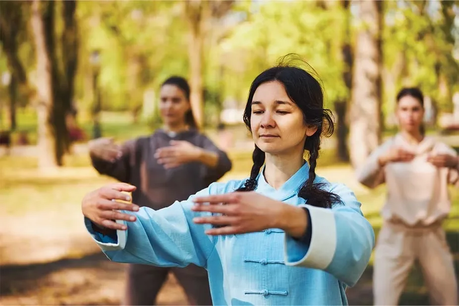 Tre donne che praticano il Tai Chi in un parco durante il giorno, con alberi e luce naturale.