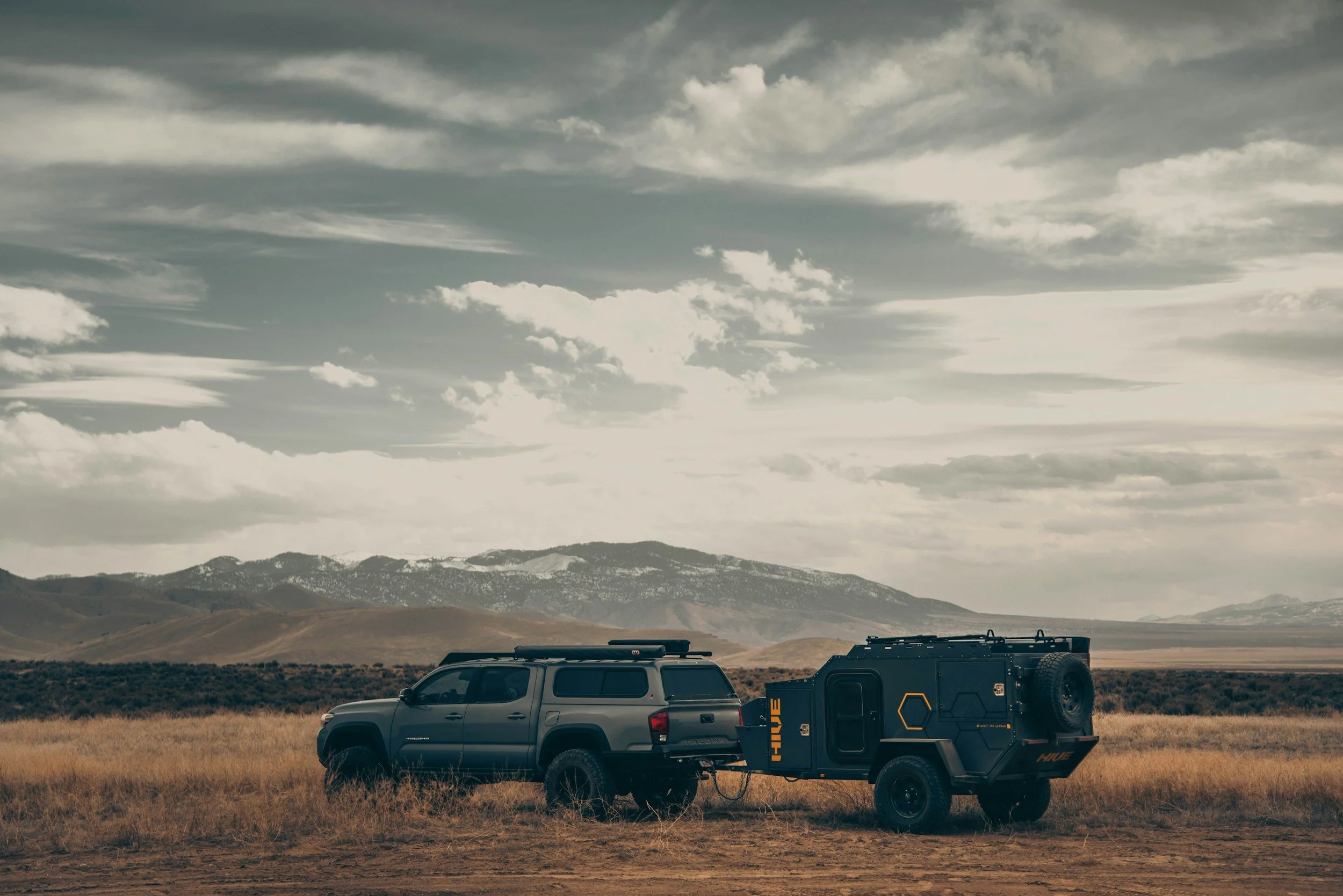 A dark-colored pickup truck towing a boxy, black off-road camper trailer in a vast open plain with mountains and a cloudy sky in the background.