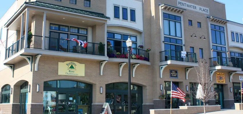 Street view of a modern brick building with balconies, signage for stores including River Outfitters and a yoga studio, American flag, street lamp, and leafless trees.
