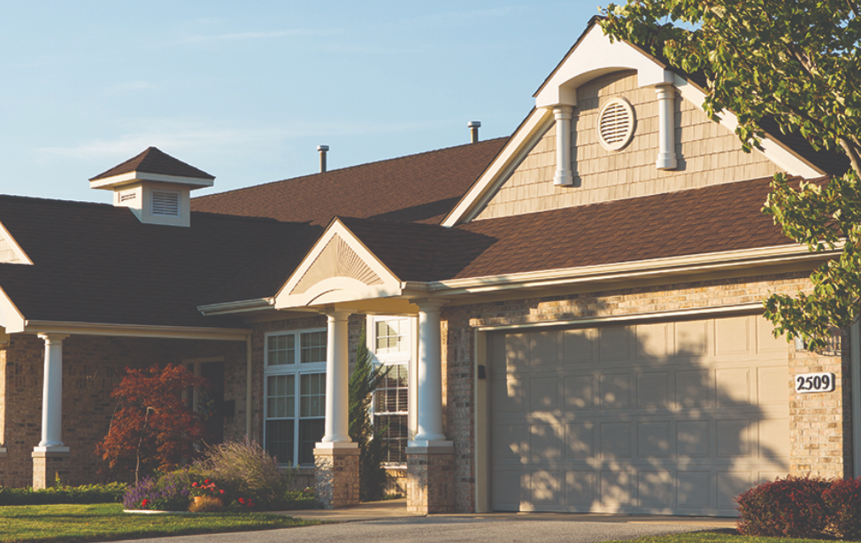 A suburban house with a brick exterior, a garage door, and a gabled roof with brown shingles, surrounded by landscaped bushes and trees during daytime.