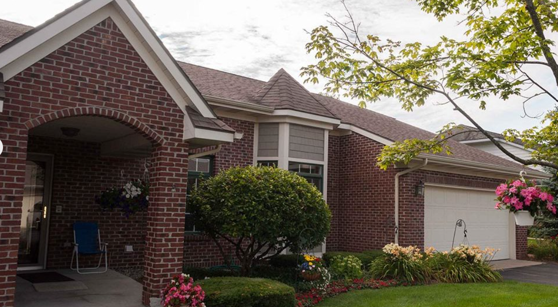 Front view of a brick house with a white garage door, garden with flowers, bushes, and tree, and a porch with a blue chair.