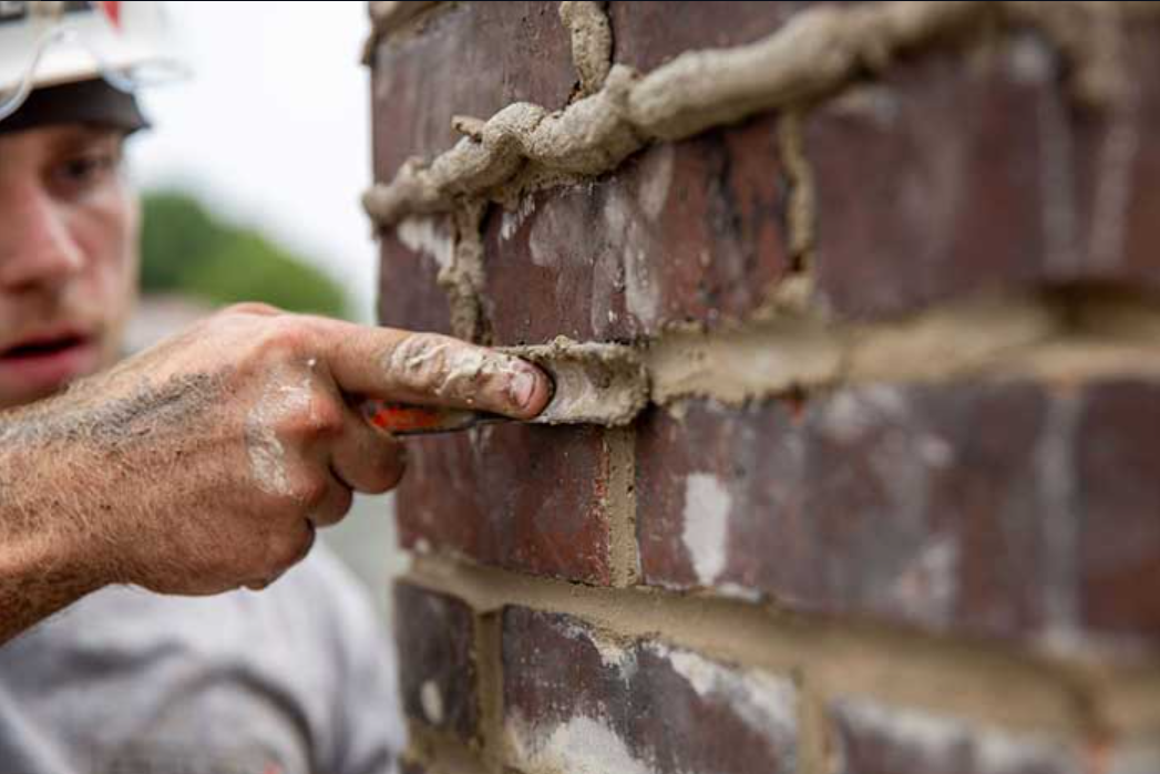 A construction worker pointing at a brick wall with a trowel, applying mortar to the bricks.