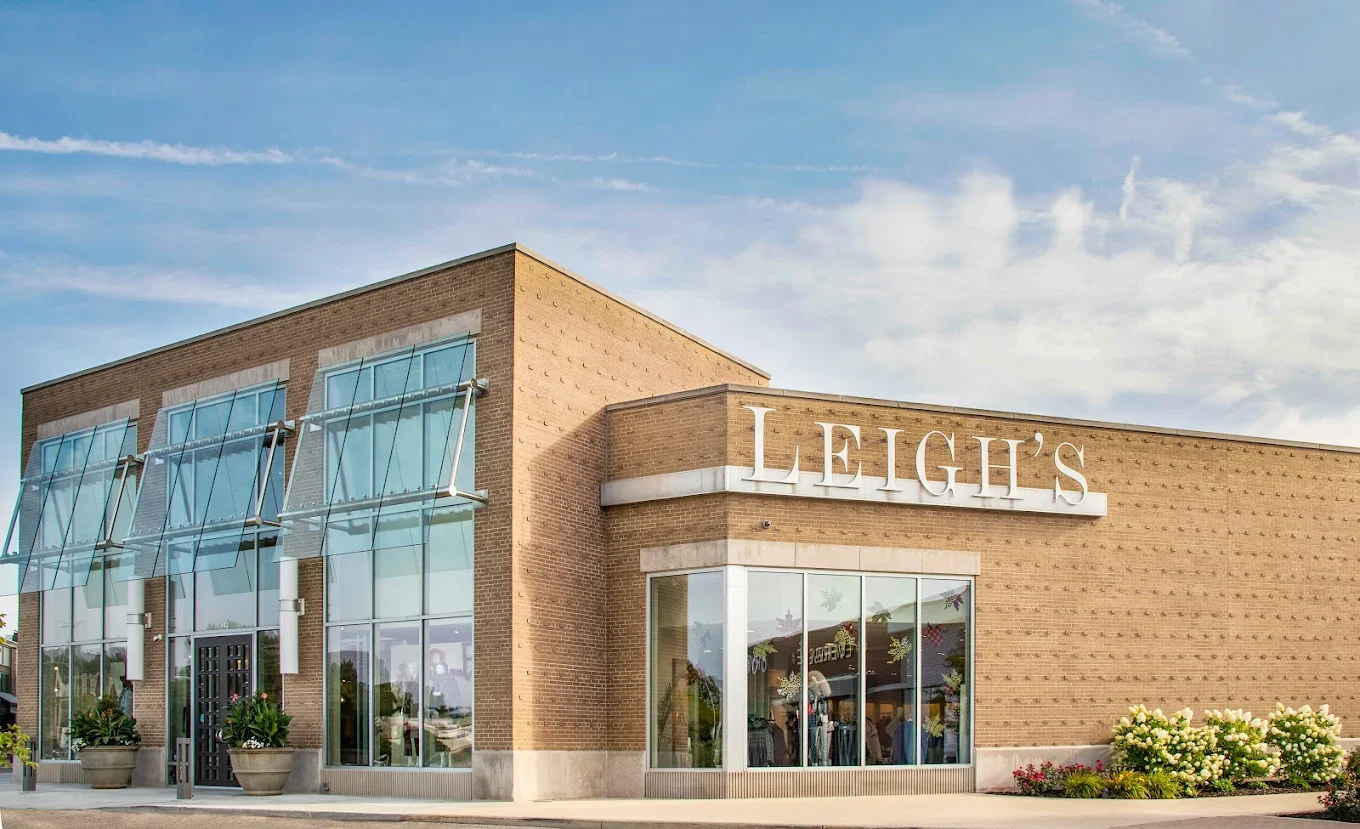Exterior view of Leigh's store with glass windows, brick facade, and a large sign displaying 'LEIGH'S' in white letters, surrounded by flowering plants and clear sky.