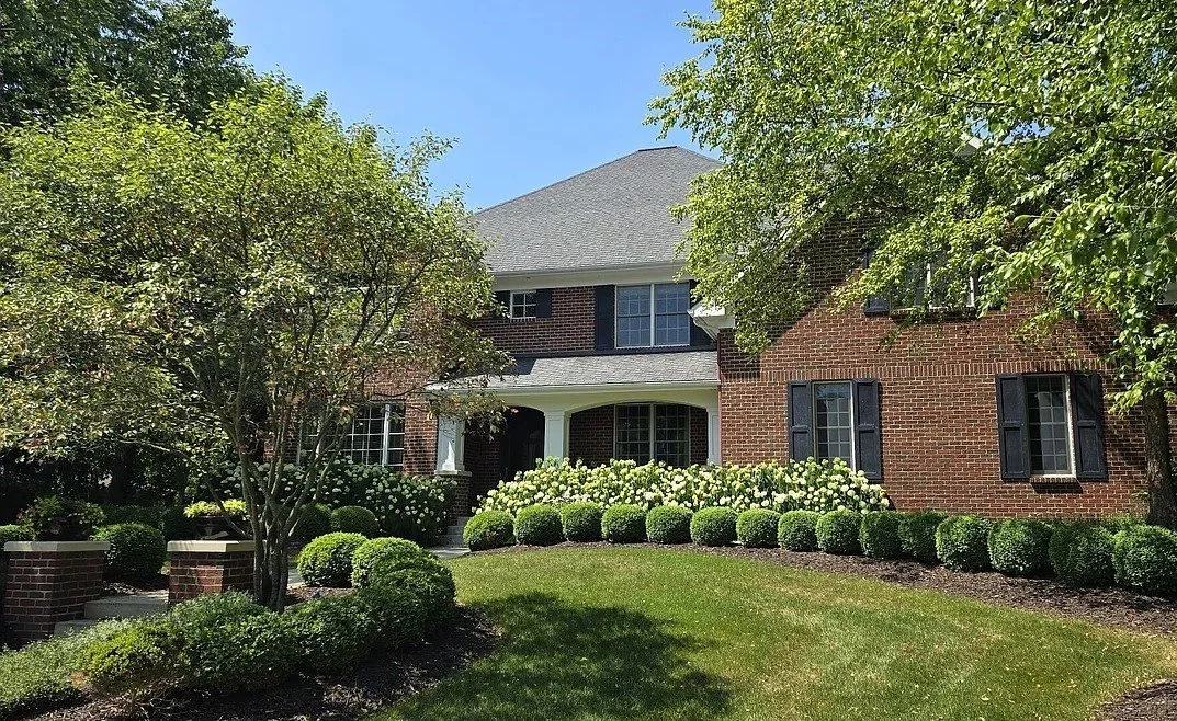A brick house with black shutters, surrounded by trimmed bushes, trees, and a green lawn under a clear blue sky.