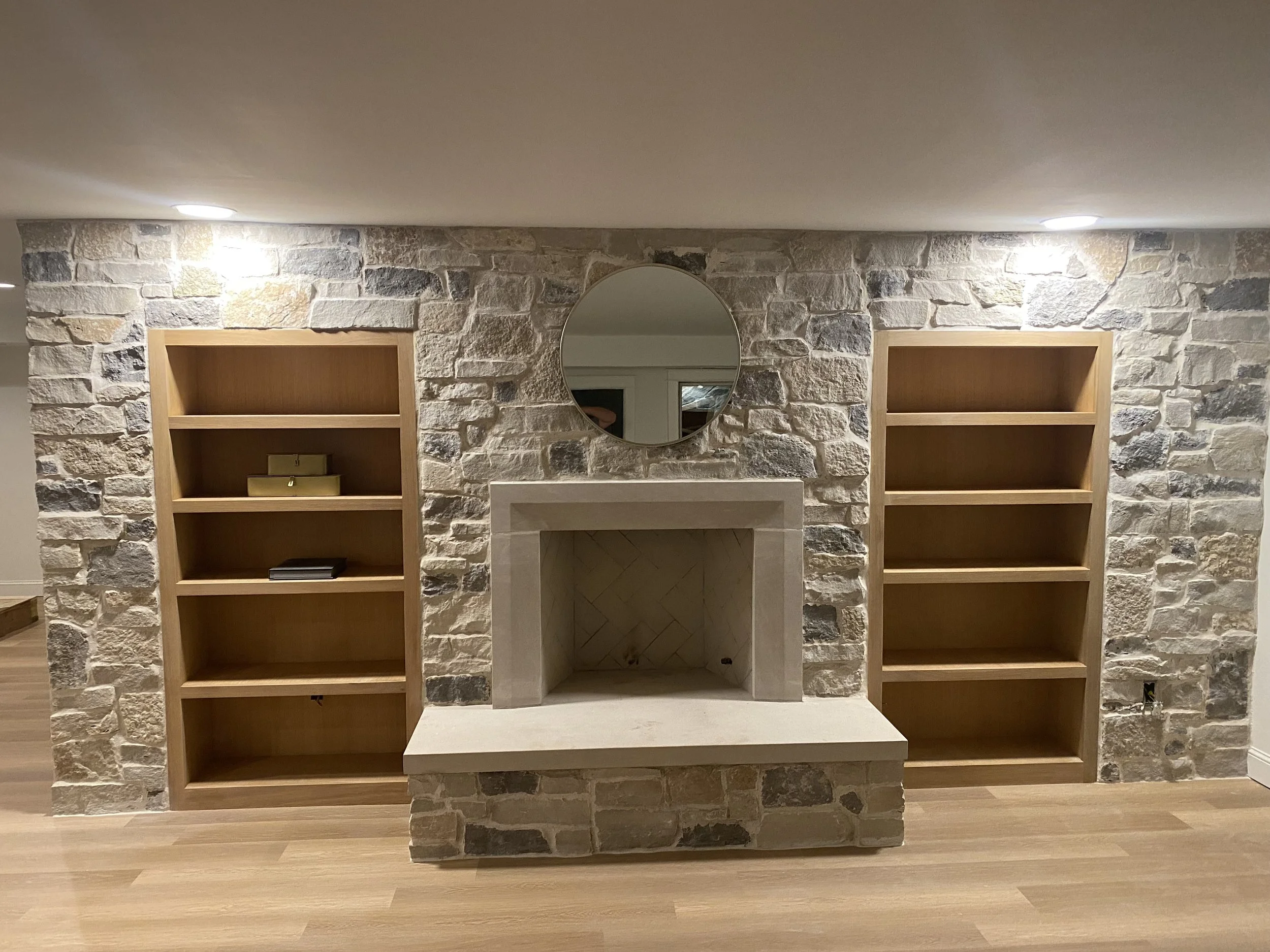 A stone fireplace with a round mirror above it, flanked by two wooden bookshelves with five shelves each, in a room with wood flooring.