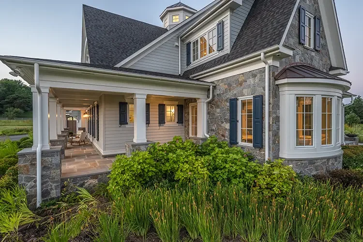 Front view of a large house with a stone and siding exterior, black shutters, a covered porch, and surrounding greenery.