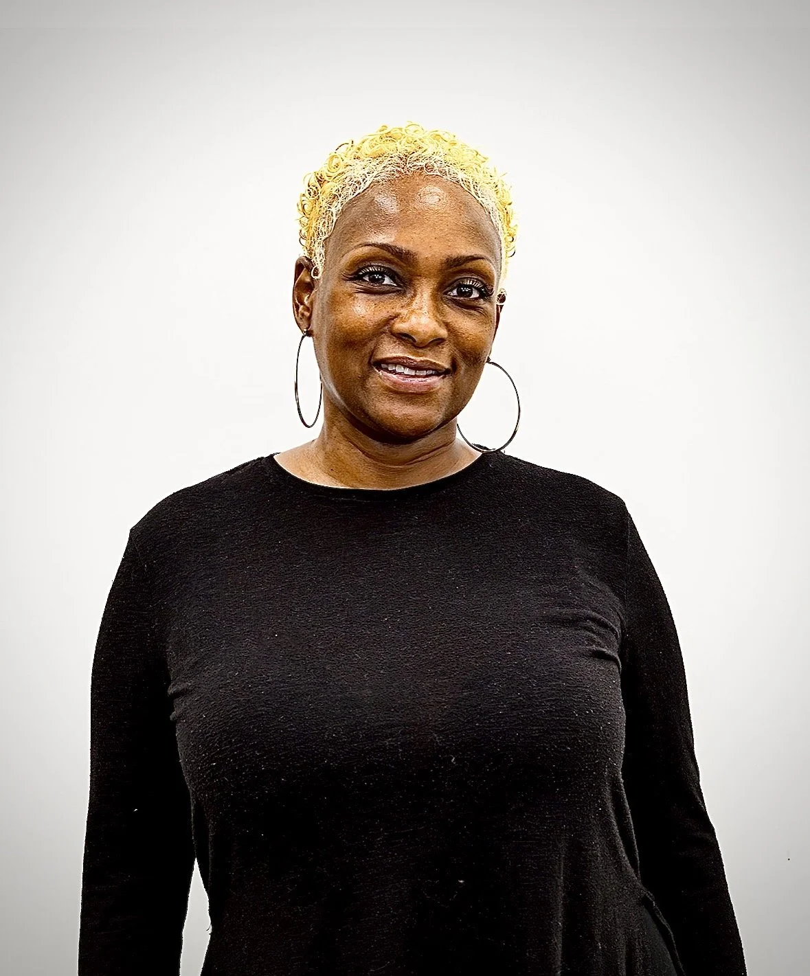 A woman with short, curly, blonde hair and hoop earrings, wearing a black long-sleeve top, standing against a plain white background.