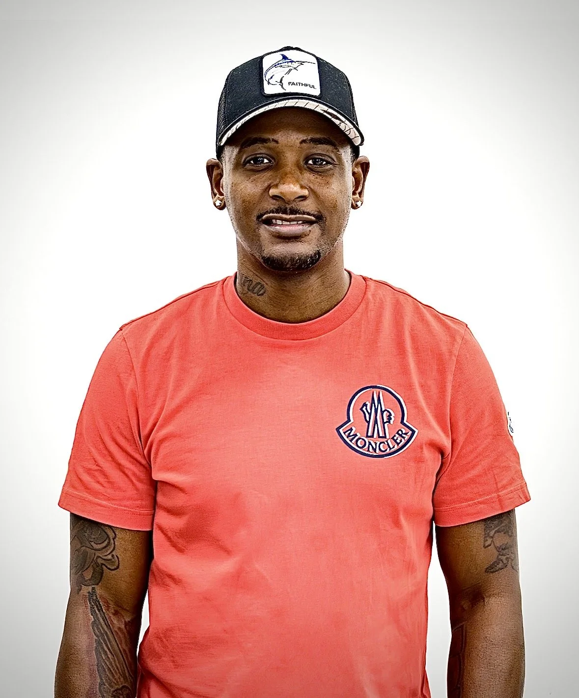A man wearing a red Moncler T-shirt and a black cap with a fish logo, standing against a plain white background.